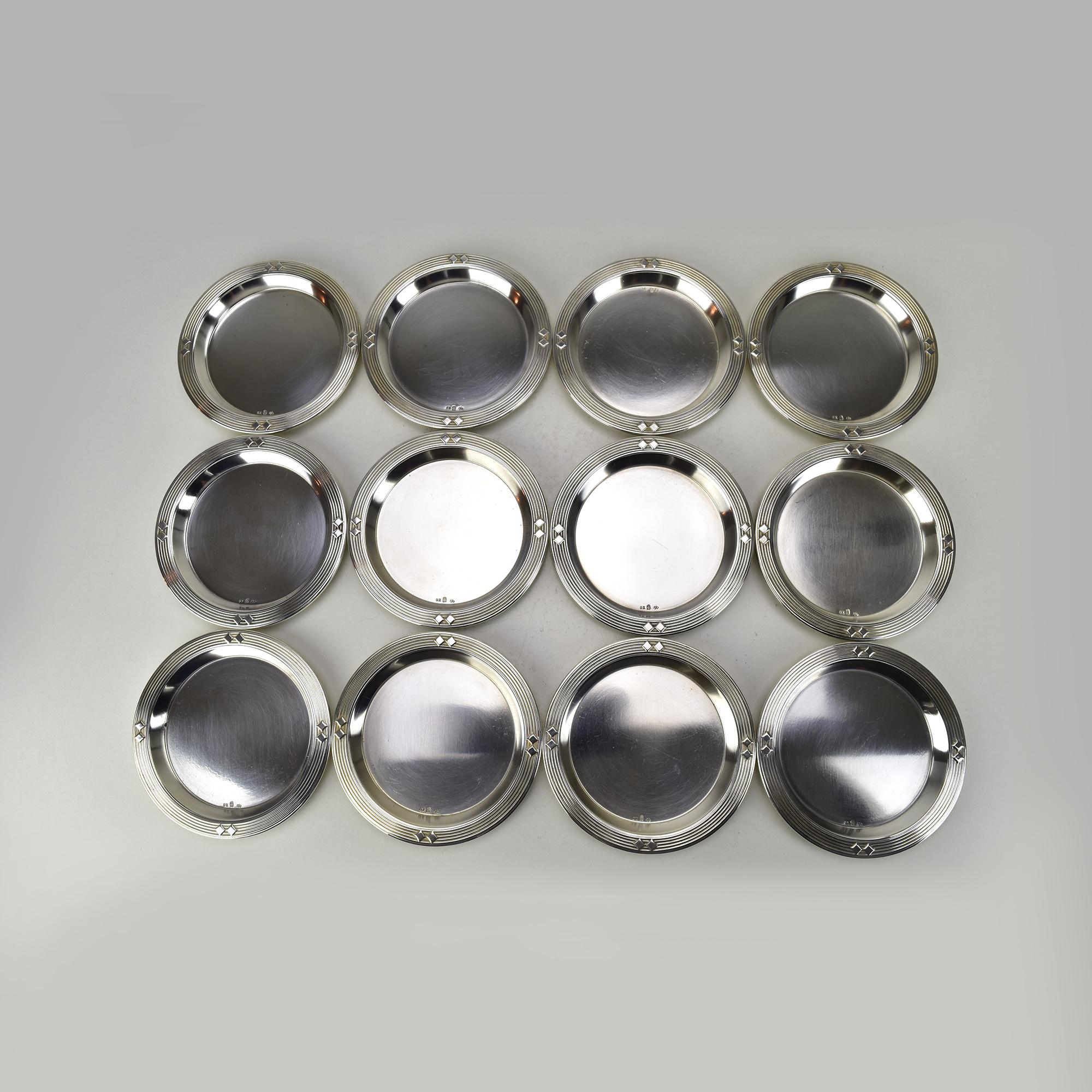 This elegant set of 14 silver-plated brass coasters was crafted by WMF (Württembergische Metallwarenfabrik) and showcases a refined Art Nouveau Secessionist design. The set includes twelve glass coasters and stwo slightly larger bottle coasters.