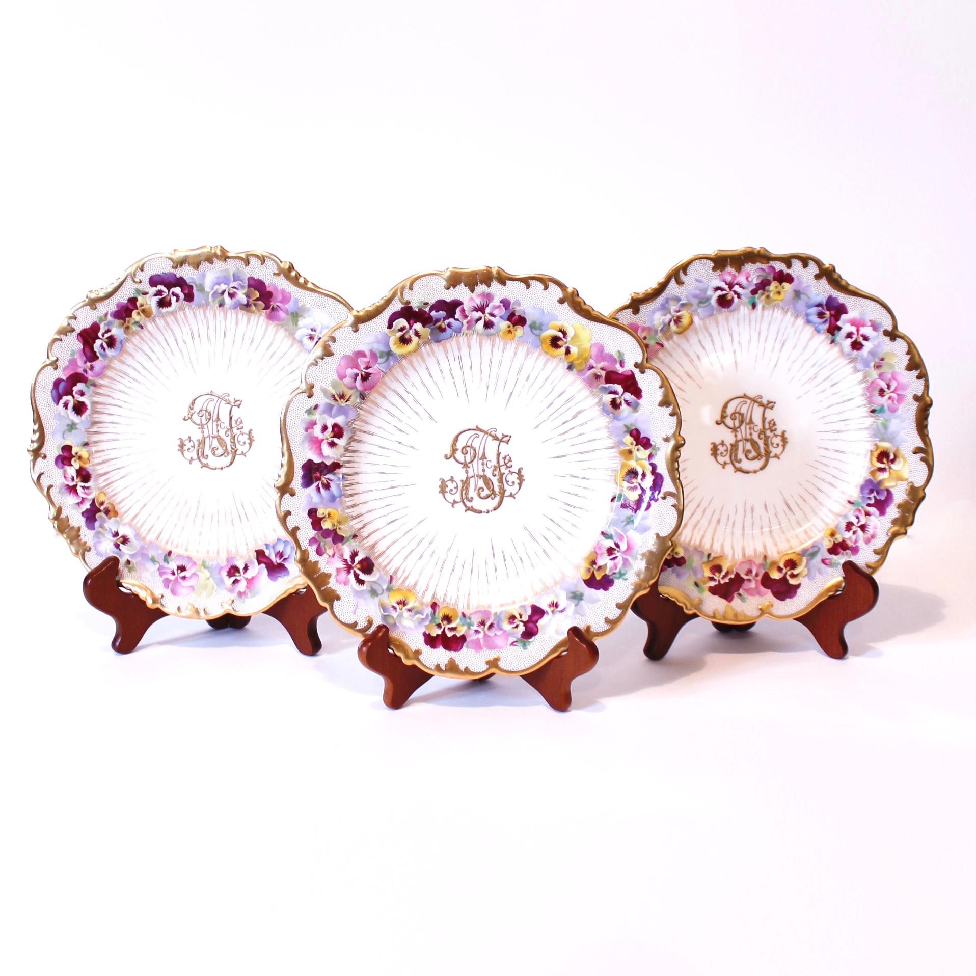 A remarkably large and beautiful set of richly gilt decorated antique English porcelain dessert plates made by Cauldon, probably dating ca. 1910. Each plate is unique with each rim hand painted with a fresh hued garland of pansies. All are in