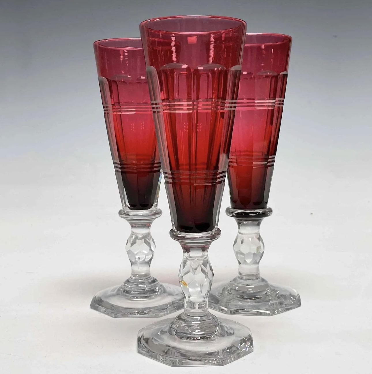 This is an amazing example of the finest hand blown crystal in the form of champagne flutes. The body is a deep cranberry that is cased in clear crystal and then decorated with an Art Deco sculptural cut decoration. The clear stem reaches down to