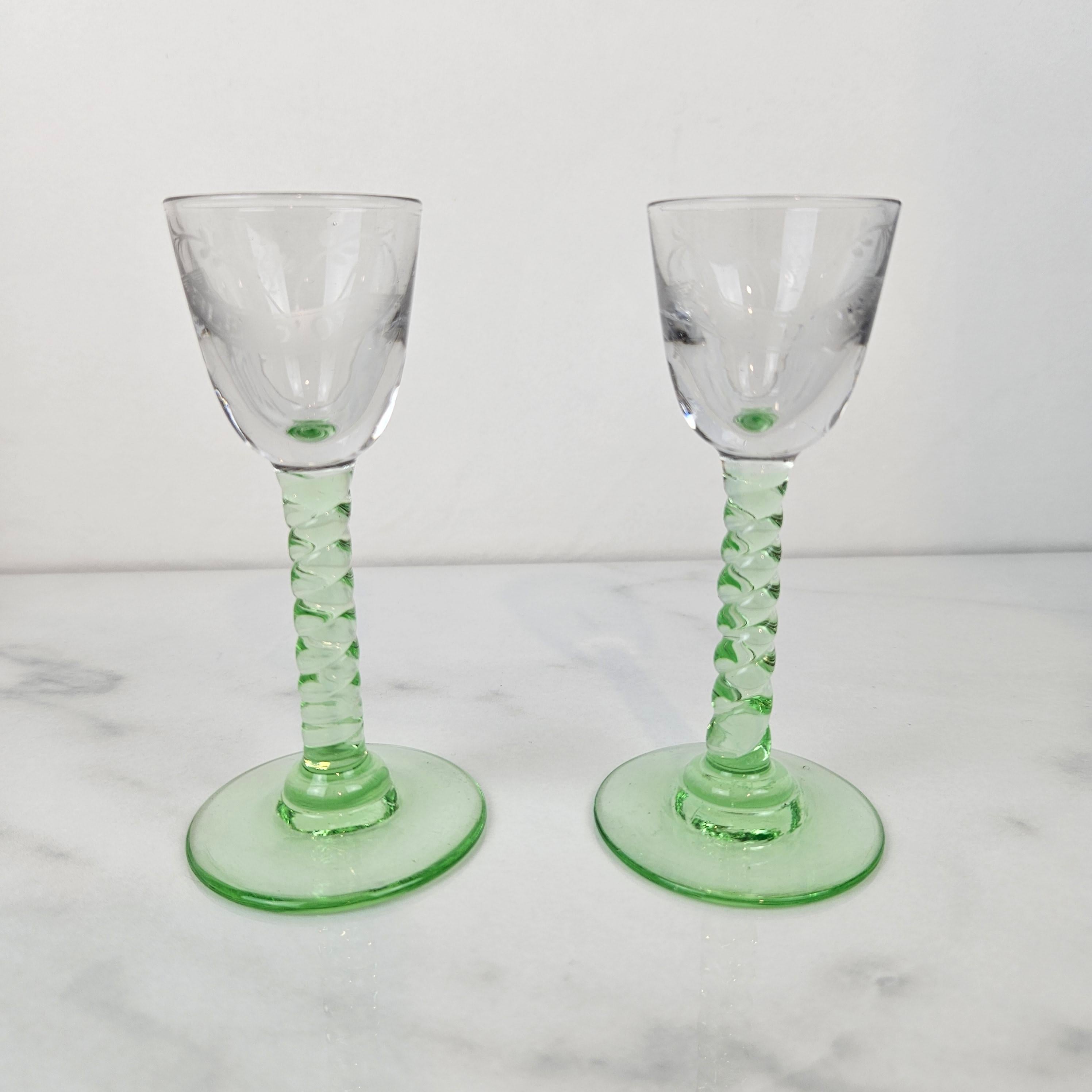 A rare and beautiful set of two antique handblown jenever glasses, engraved with the inscription “Blé d’Or.” 
Dating from around 1900, these elegant glasses feature clear bowls delicately etched with text and slender green stems with a twisted