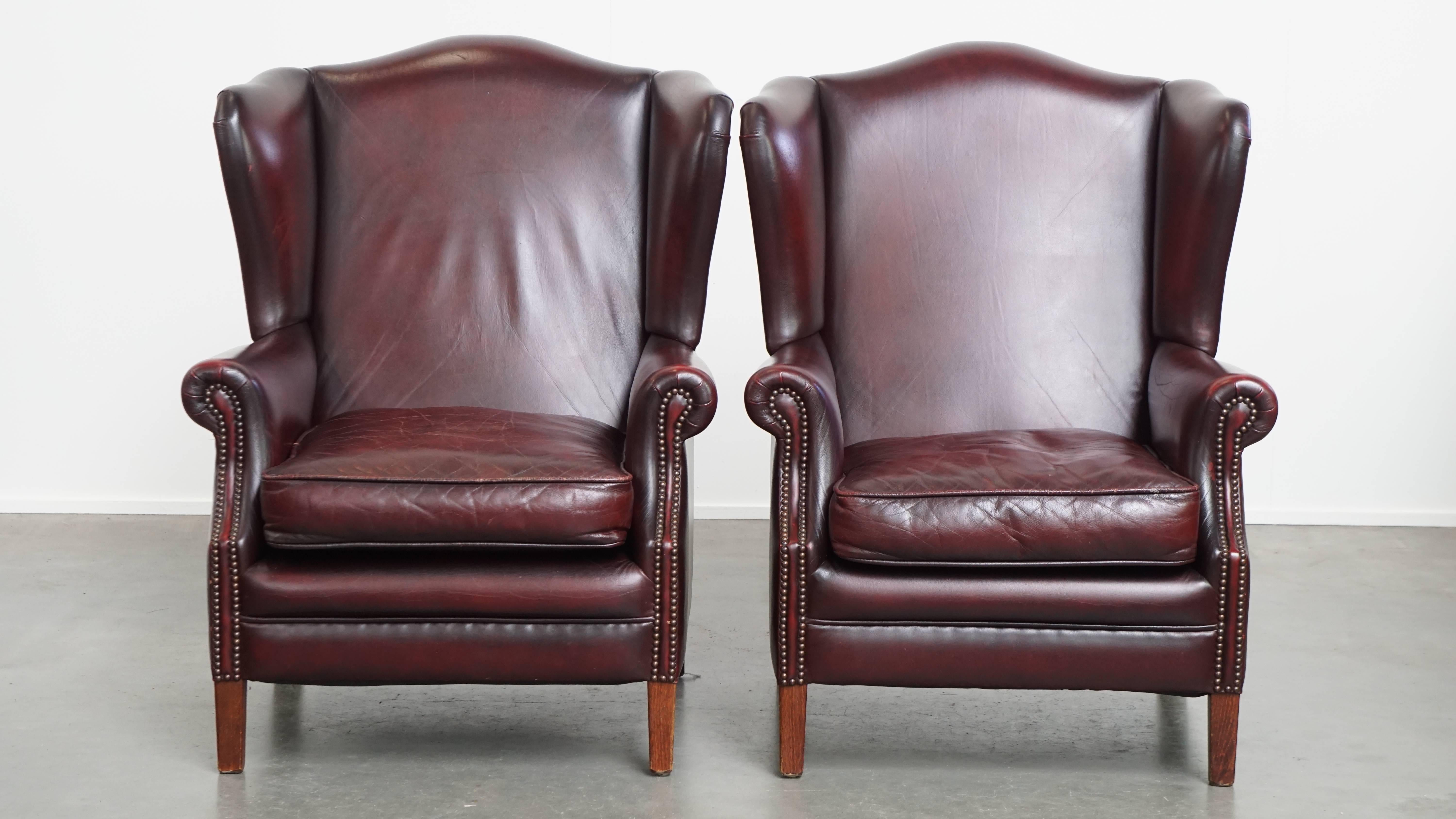 Offered: this luxurious set of two comfortable burgundy leather wingback armchairs with matching ottoman in English style, featuring a stunning patina. Made from high-quality cowhide leather and finished with decorative studs, these chairs exude