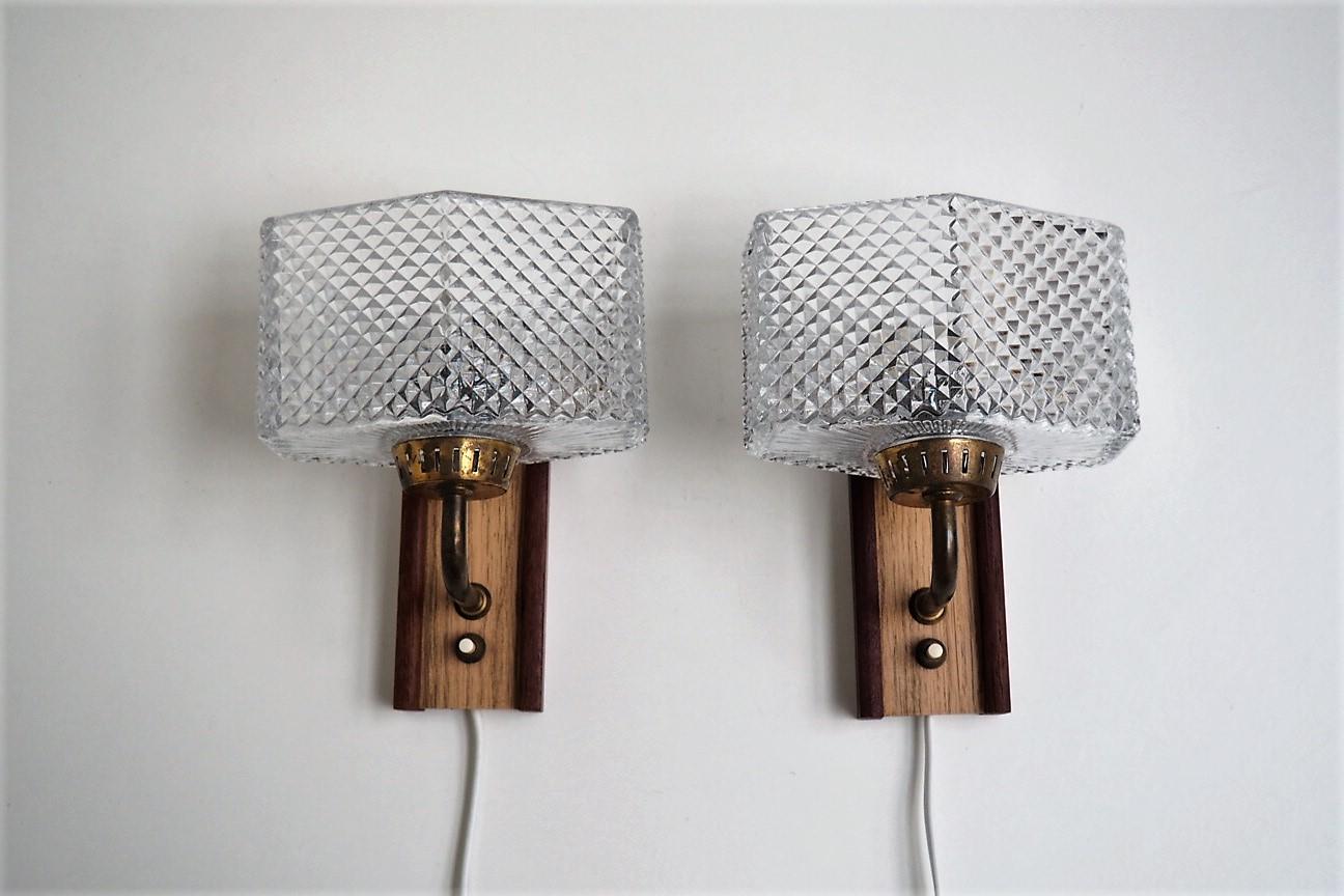Set of 2 wall sconces with wall base made in a mix of teak and foil coated metal (wood look). The shape of the shades is very special and they are made in clear glass with a beautiful molded pattern that reflectes the light very beautiful.

The