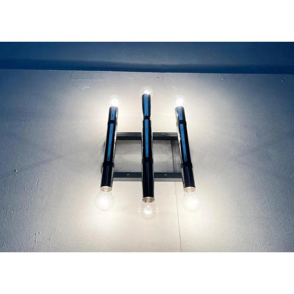 Due imponenti lampade da parete dell'era spaziale.

Un design minimalista. Tre cilindri cromati che corrono in parallelo sono sostenuti da un semplice telaio in acciaio. La lunghezza del cilindro diminuisce verso l'esterno. All'estremità superiore e
