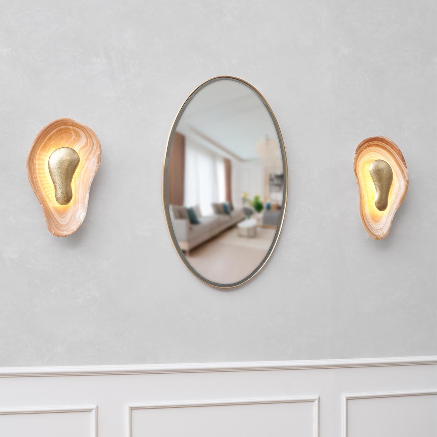 La lampada da parete Oyster presenta due sorprendenti forme speculari, meticolosamente realizzate in raro onice arancione, ognuna delle quali presenta venature uniche che ne esaltano la bellezza naturale. Il centro, un elemento in ottone martellato,