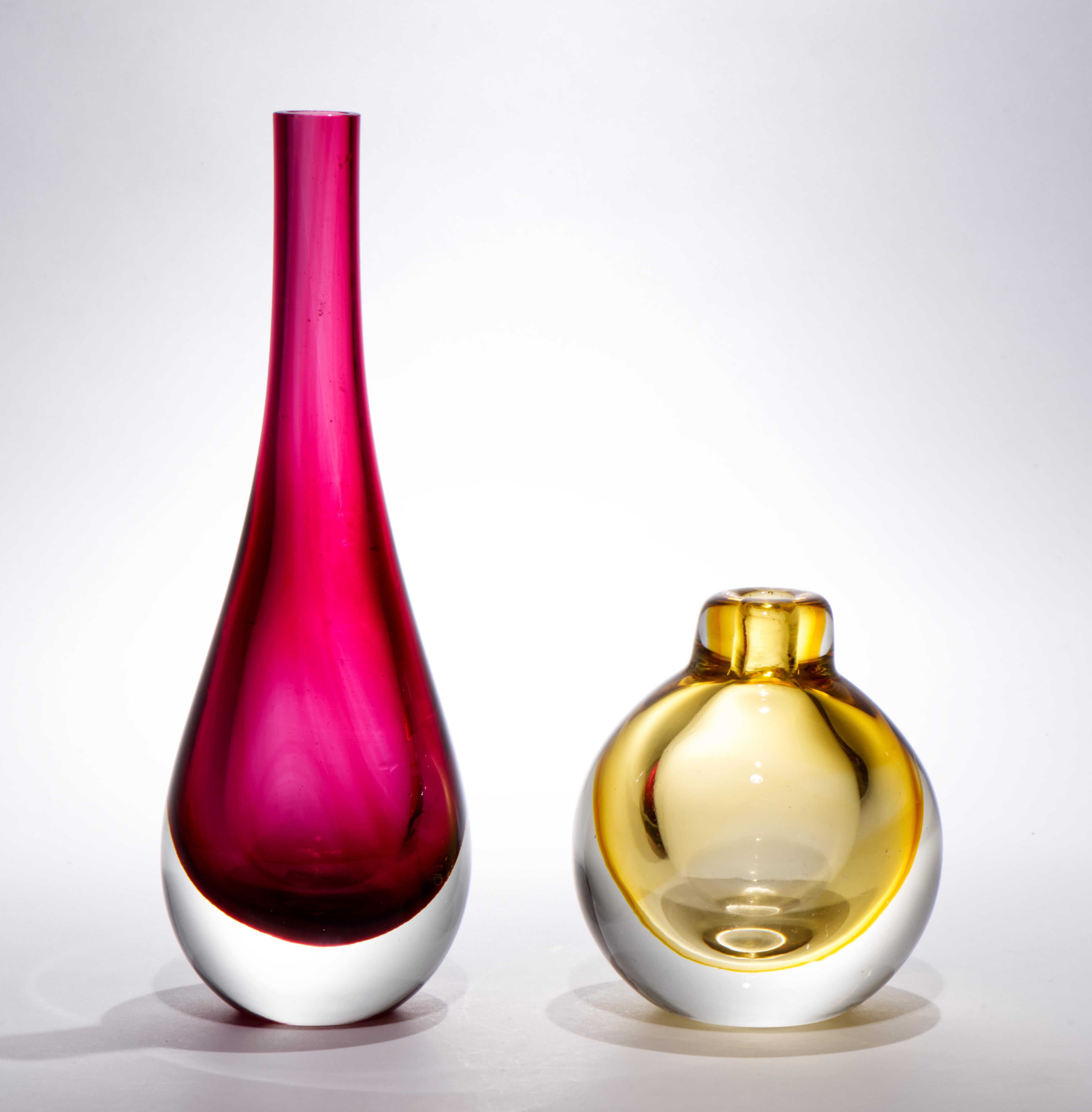 Das Set aus 2 Knospenvasen aus Vintage-Kunstglas zeichnet sich durch weiche, abgerundete Formen und eine zurückhaltende Farbpalette aus. Die größere Vase ist aus cranberryrotem Glas, die kleinere aus bernsteingelbem Glas; das farbige Glas ist in