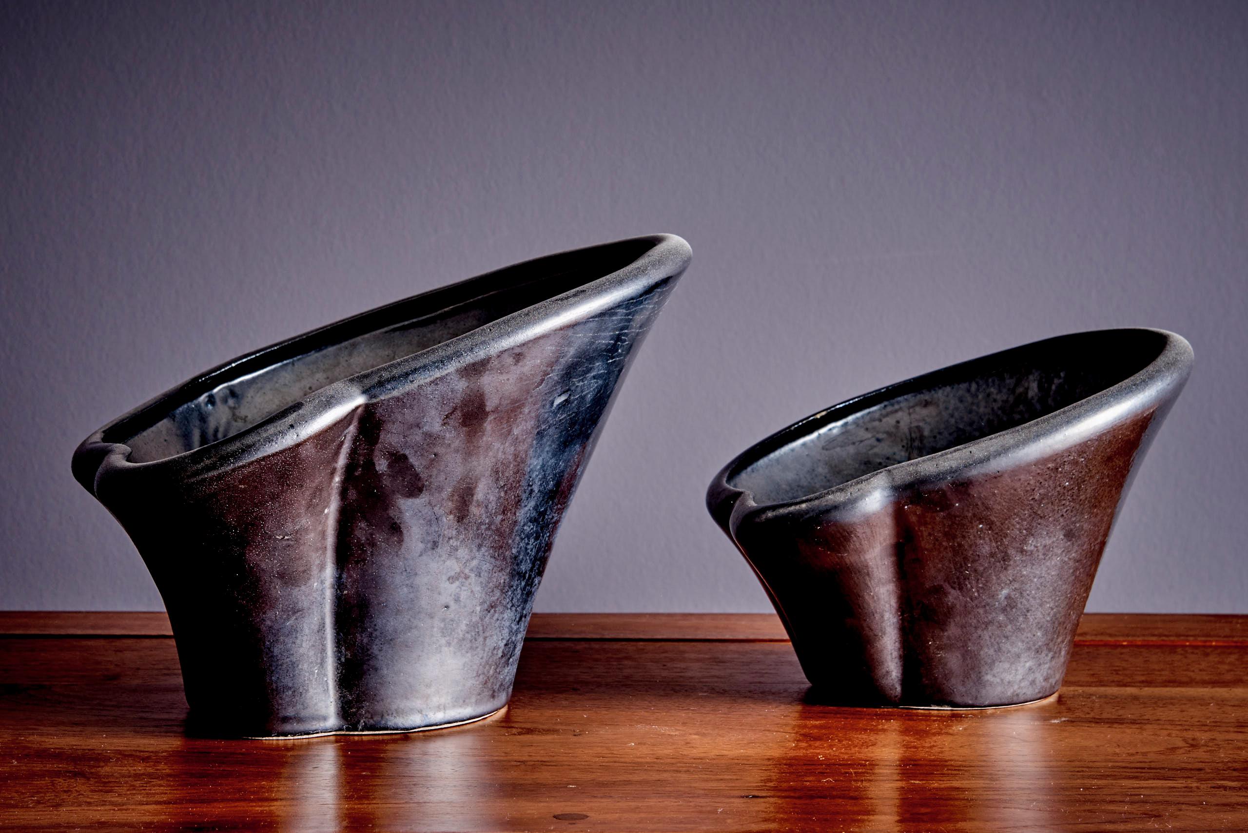 Pair of handmade ceramic planters with a unique sculptural shape and dark metallic glaze. Each bowl has an organic, flowing form and a textured surface with natural variations. The larger one has a height of 20cm and a diameter of 30 cm. The smaller