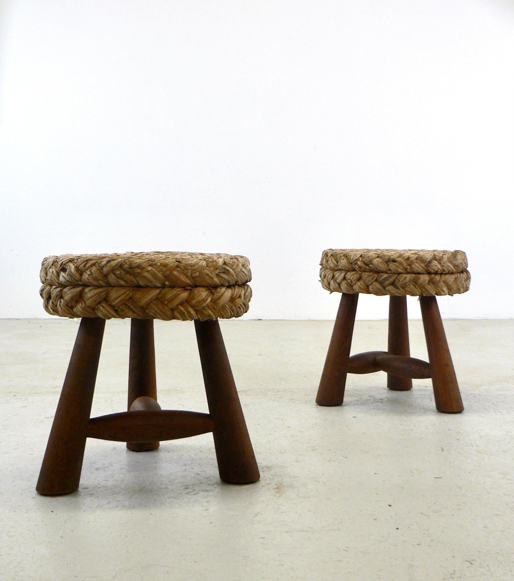 Mid-century French low Shepard's stools designed by Adrien Audoux and Frida Minet. This rustic French poufs showcase a circular seat crafted from woven raffia/rattan, supported by tripod legs and an oak keel base. Maintaining its original condition.