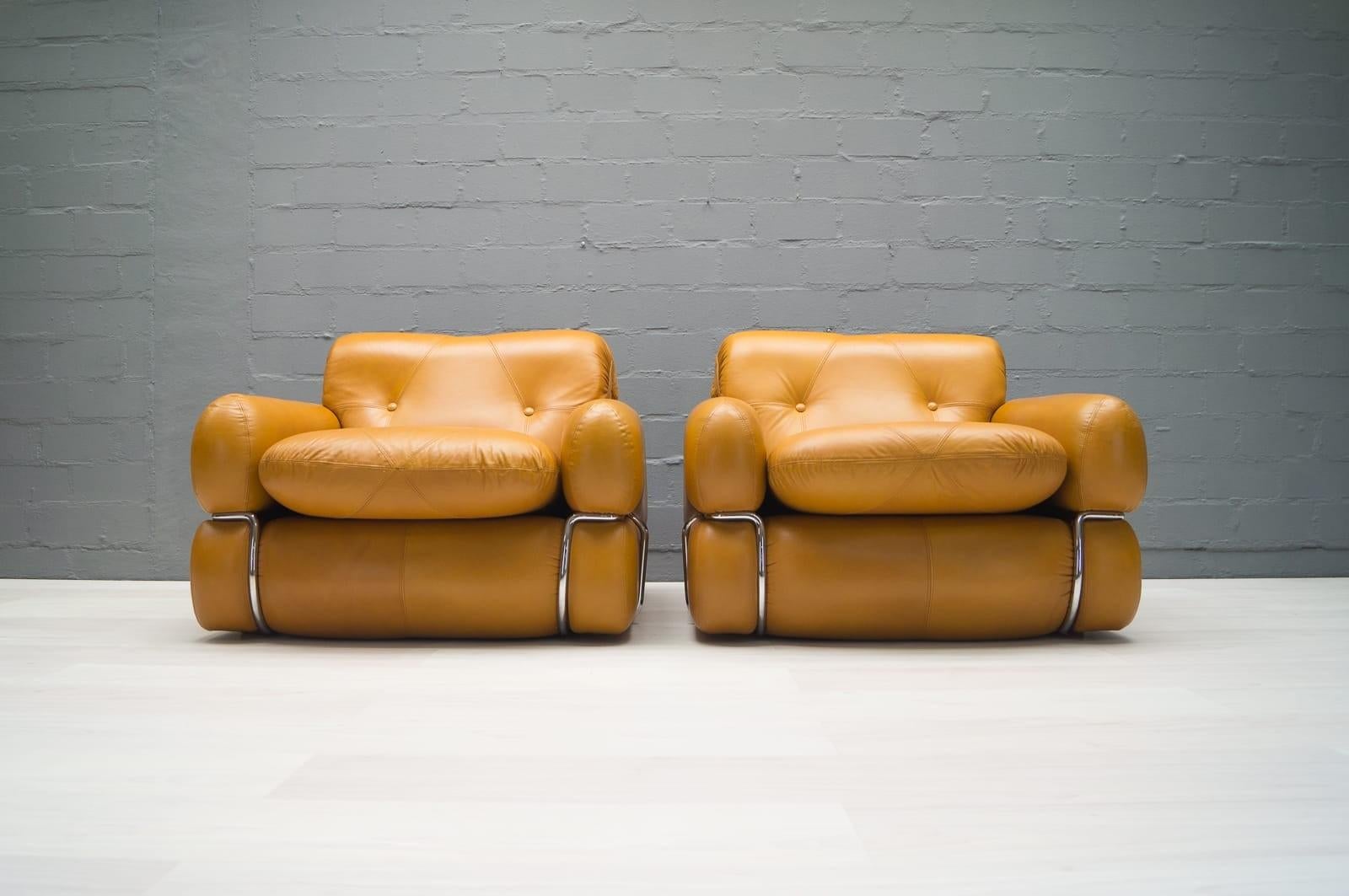 This pair of freestanding armchairs are covered in cognac colored leather and sit on chromed metal frames. 
The seat height is 40cm, the depth of the seat is 52cm, the width is 60cm.

 