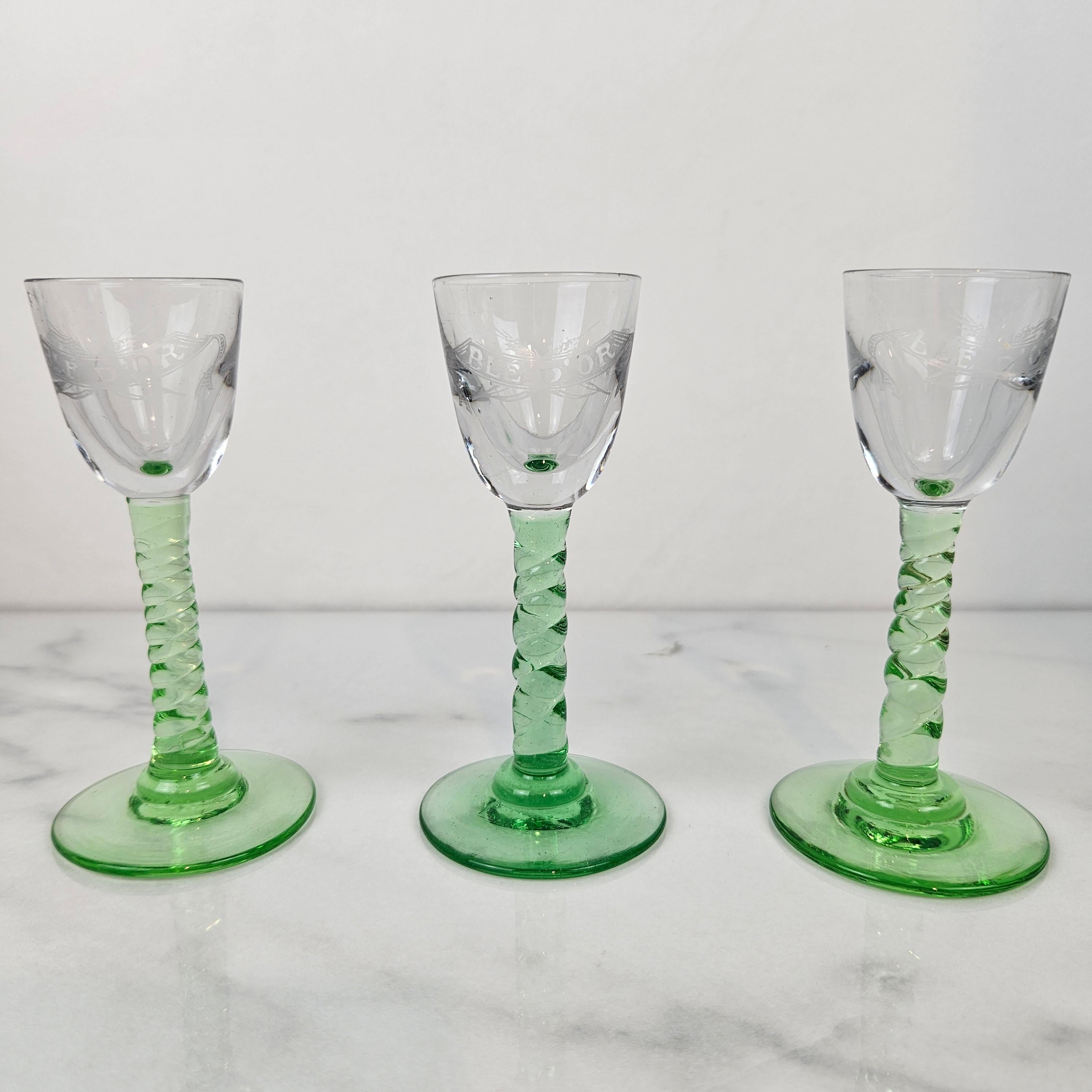 A rare and beautiful set of three antique handblown jenever glasses, engraved with the inscription “Blé d’Or.” 
Dating from around 1900, these elegant glasses feature clear bowls delicately etched with text and slender green stems with a twisted