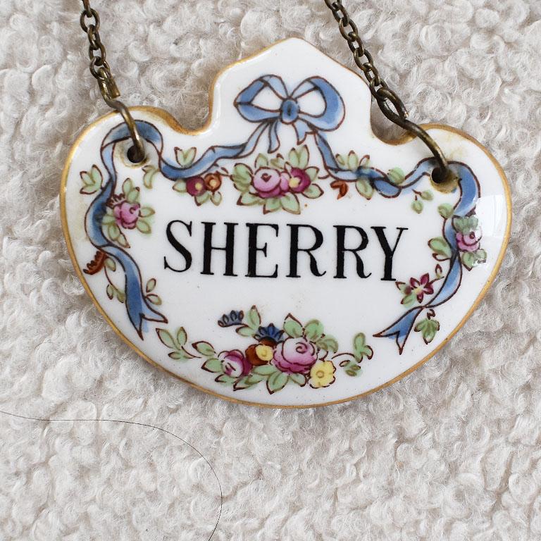 A set of three antique liquor bottle decanter tags. Each tag is created from fine porcelain in a medallion shape. On a crisp white background, each is hand-painted with a blue, pink, and green floral motif. A hand-painted gold edge decorates the