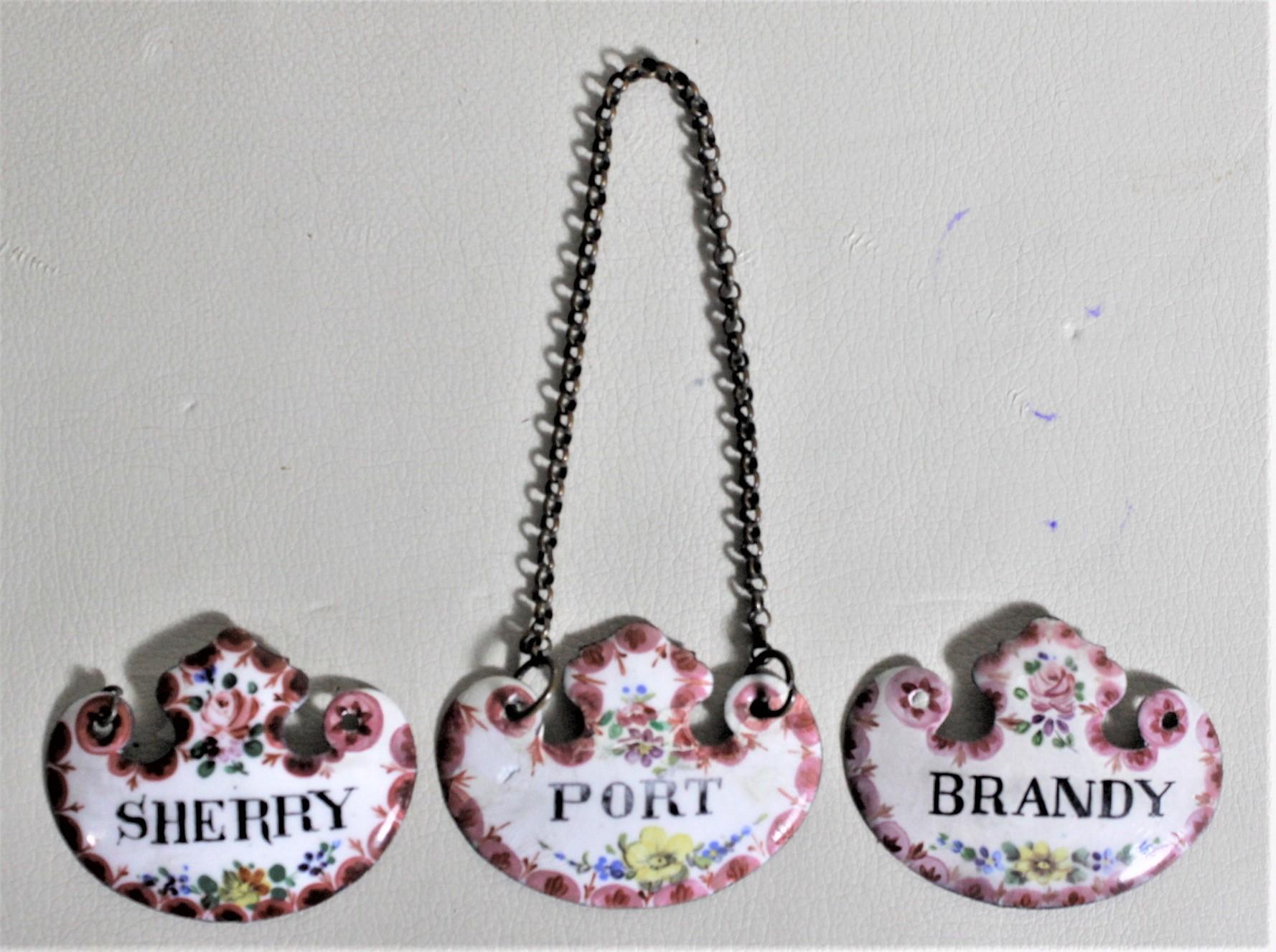 This set of three enameled liquor bottle tags are unsigned, but believed to have been made in the United States in circa 1940 in the Victorian style. The tags are done in a shield shape of enameled metal with hand-painted flowers bordering the label