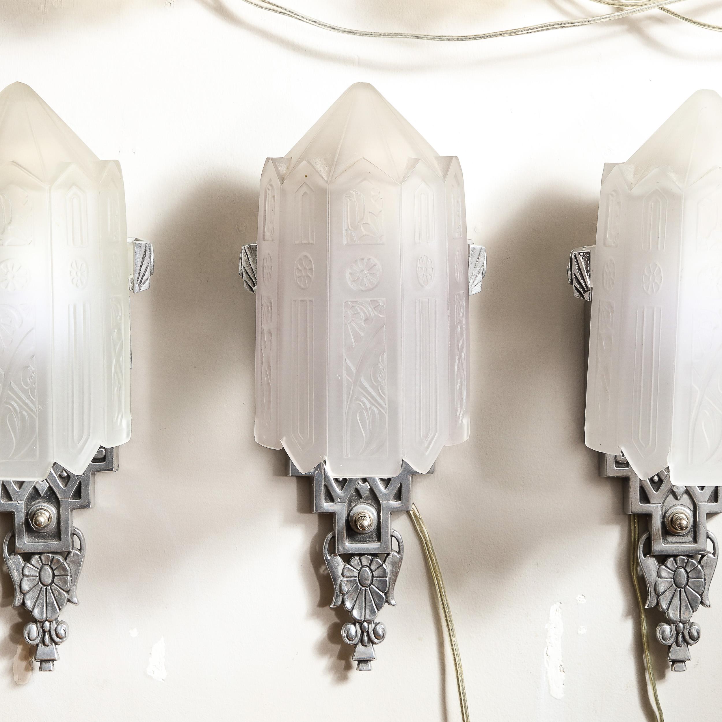 This stunning set of three Art Deco sconces were realized in the United States circa 1930. They feature skyscraper style bases in forged aluminum features scroll form detailing bookending a stacked totemic form below an abstracted striated ovoid