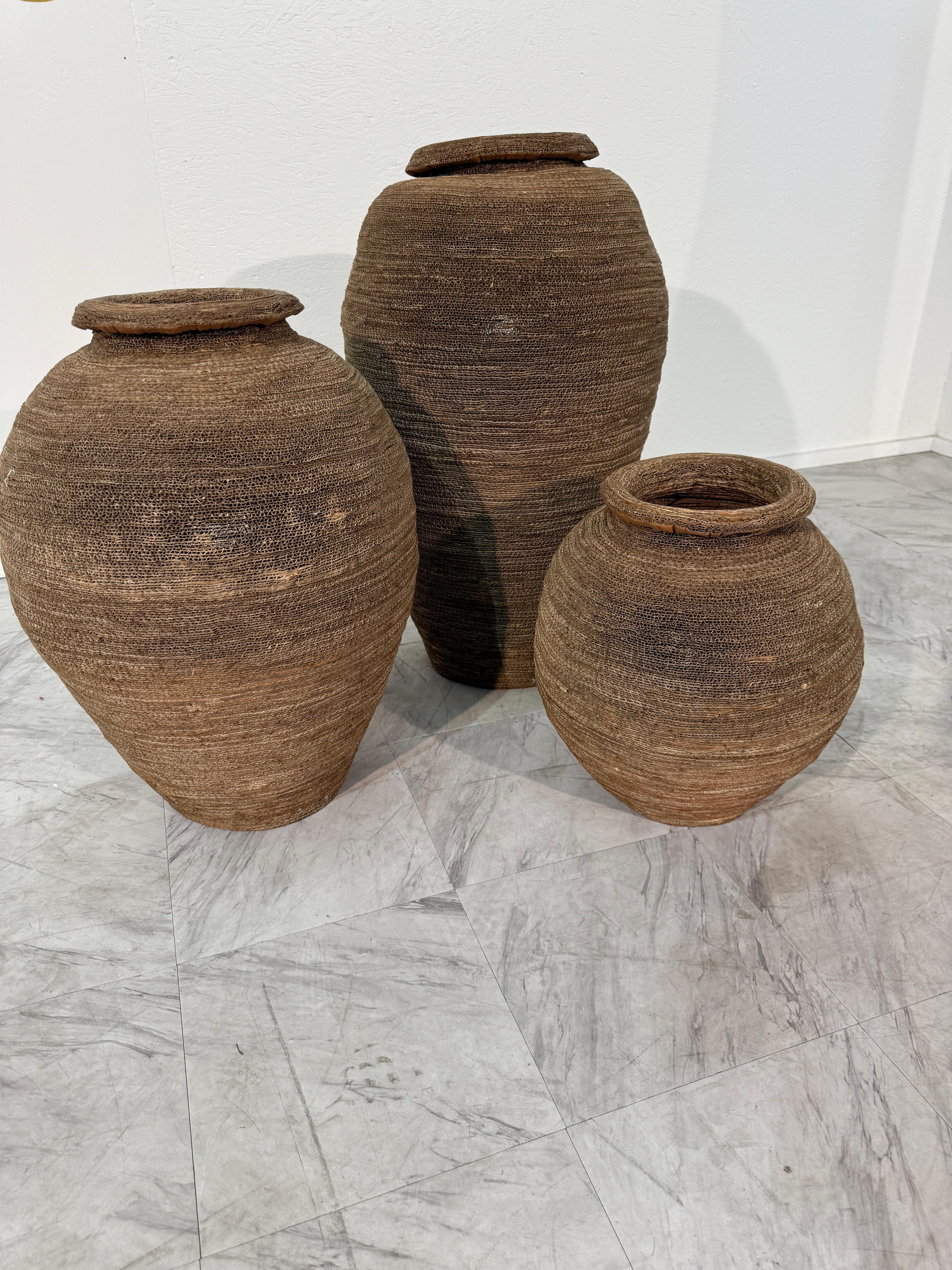 This set of three floor lamps is crafted from tightly hand-woven, coiled, and layered corrugated cardboard. Each form recalls the silhouette of an ancient amphora or woven vessel, offering both sculptural presence and organic warmth. The natural