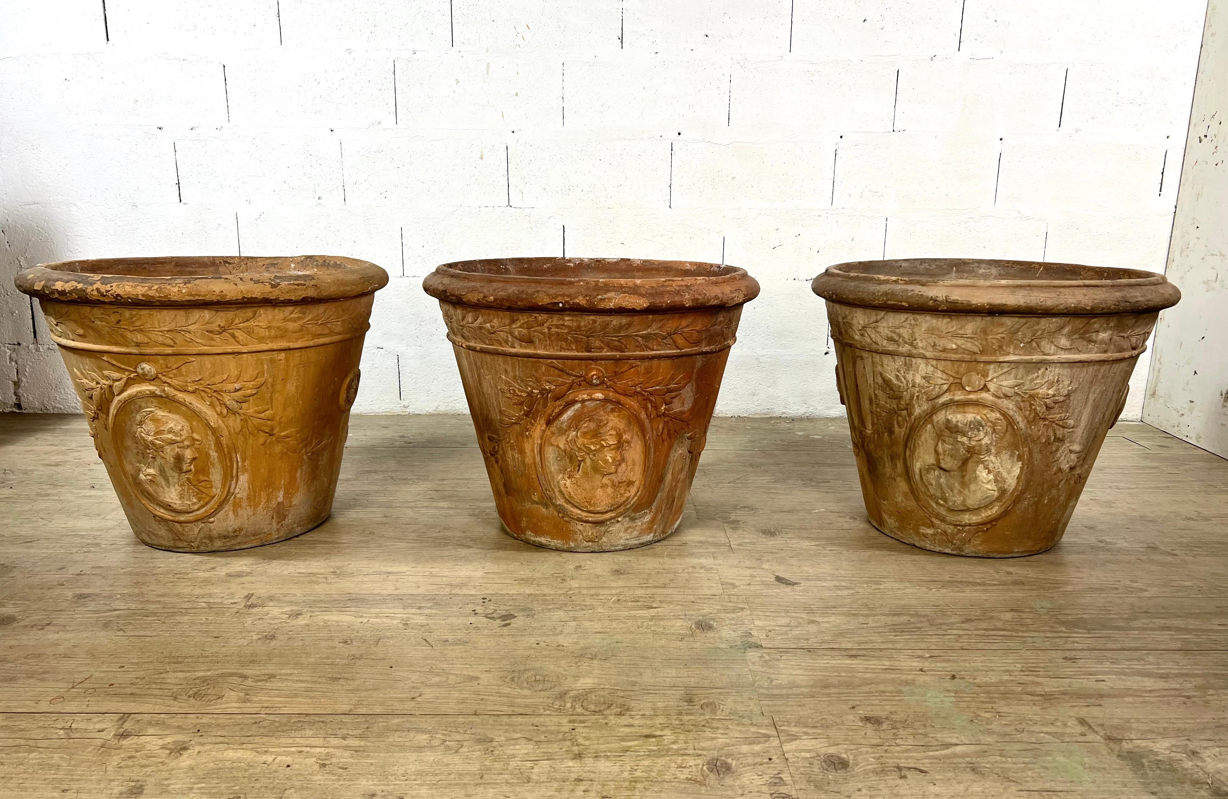 This rare set of three large patinated terracotta urns, crafted in the early 20th century, is directly inspired by the elegant aesthetics of 18th-century classical garden design. Each urn features a raised medallion with a female profile,