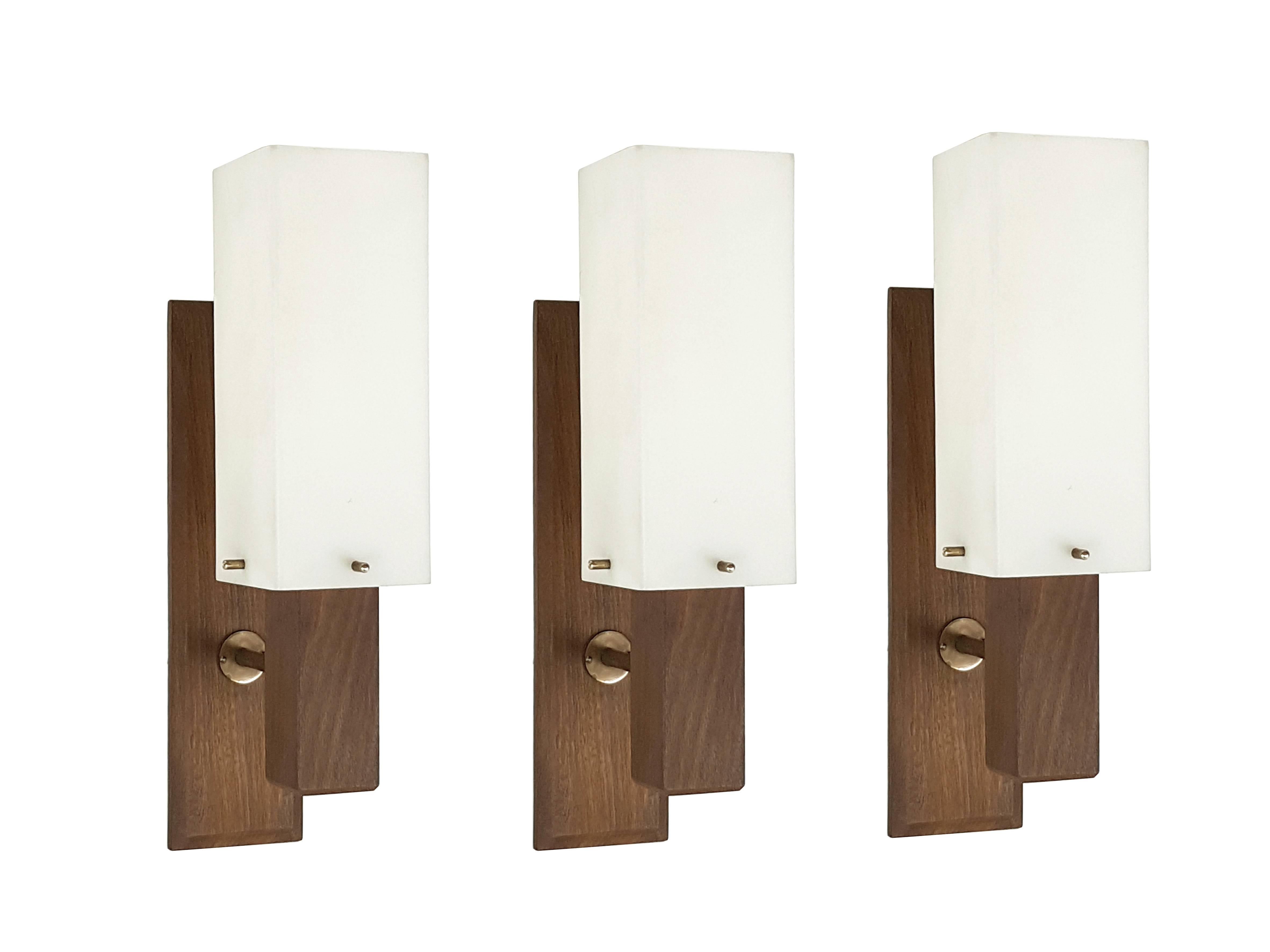 This set is composed by three wall lights made from a brass and teak structure with an opaline perspex shade. They remain in an overall very good condition. Measures: Overall height 36 cm, only shade: 21 H x 8 W x 8 D.