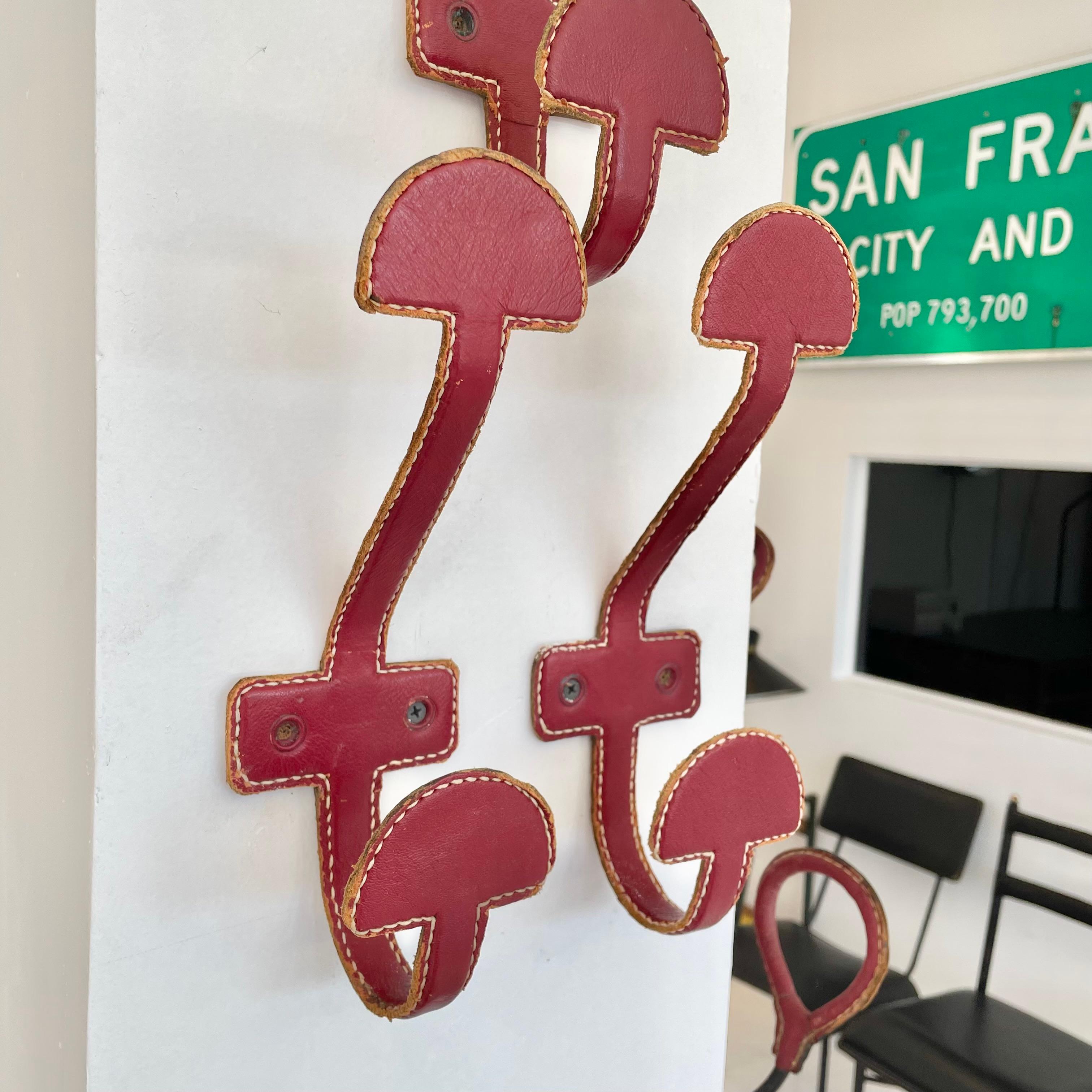 Handsome wall hooks by French designer Jacques Adnet in a beautiful red leather with perfect patina. Two places for hanging items on each hook. Frame made of solid iron and completely wrapped in leather. Every seam is stitched with Adnet's signature