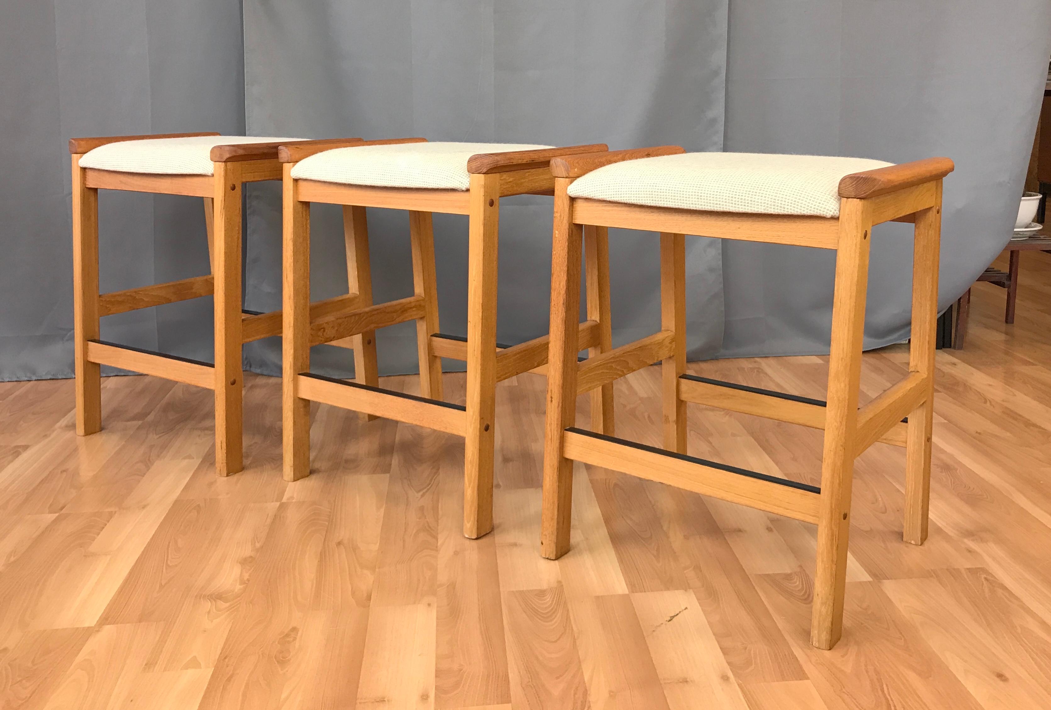 Offered here are a set of three counter stools, a J.L. Møller design for Højbjerg of Denmark.
Solid Teak legs and arms, foot rests on two sides of the stools, wool fabric upholstery in an oatmeal color.