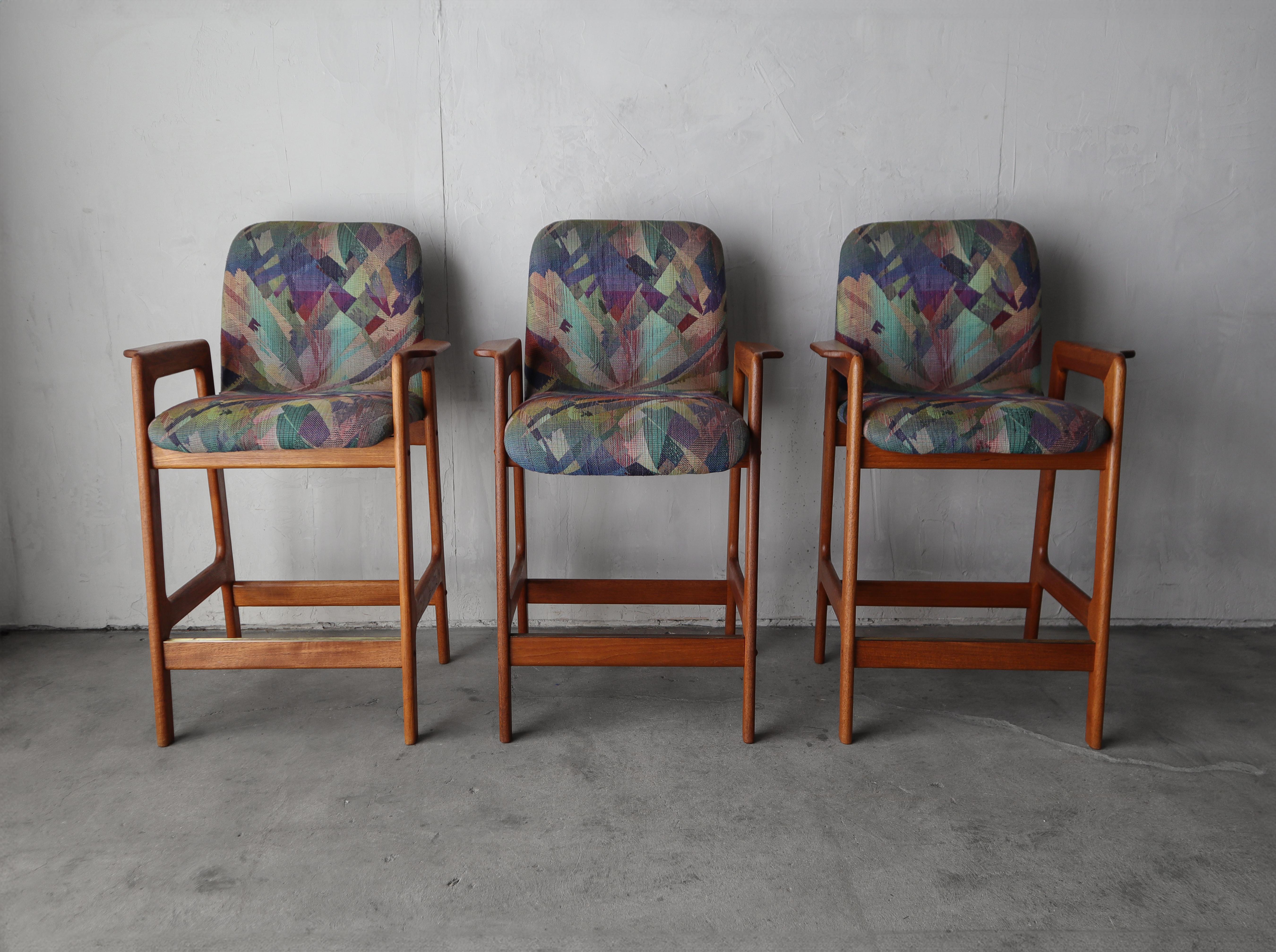 Joli ensemble de 3 tabourets de comptoir en teck danois, j'adore le fait qu'ils aient des accoudoirs et le bord en laiton sur le repose-pieds.

Les tabourets sont en très bon état avec des signes minimes d'utilisation, la seule chose notable est une