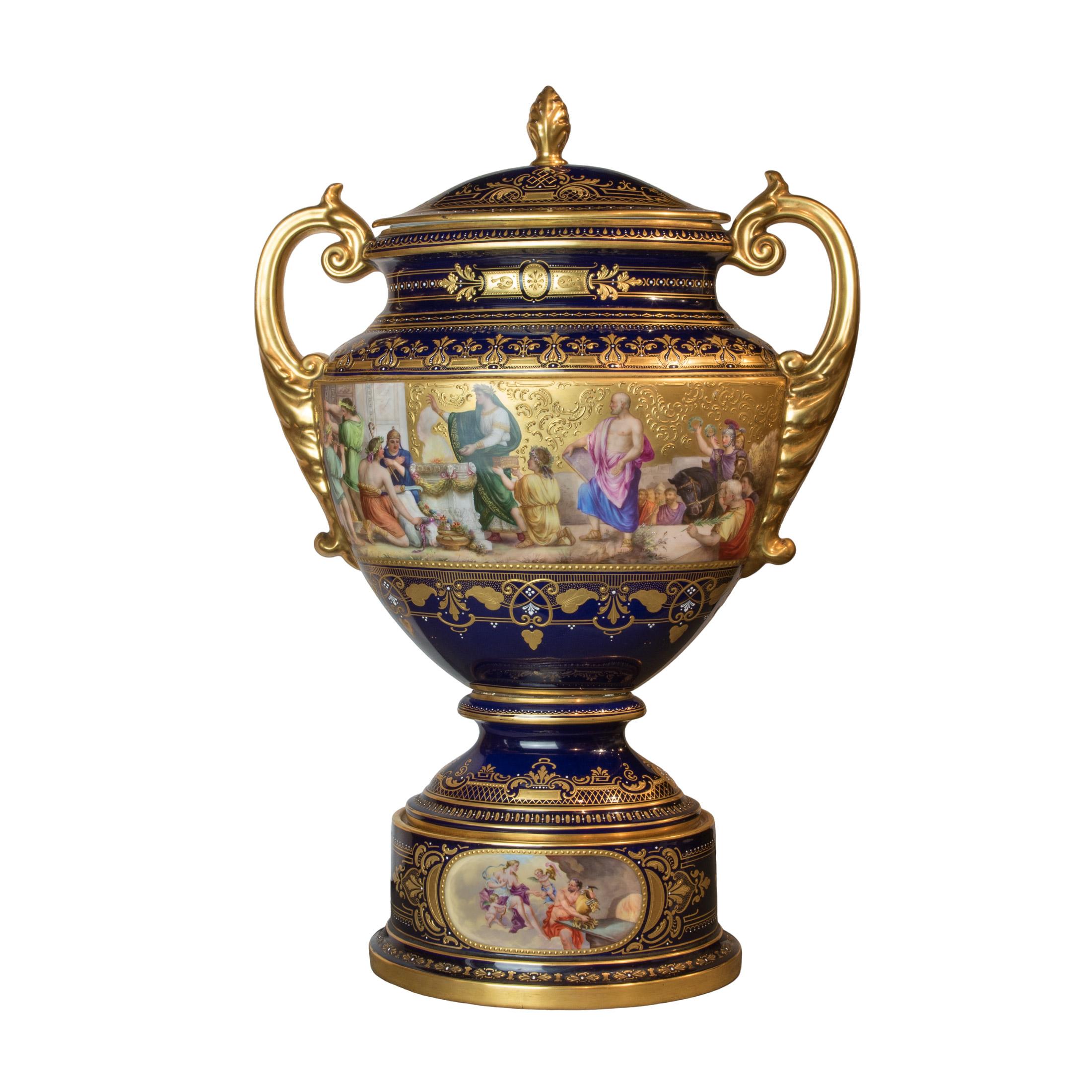 The large urn, signed by artist W. Pfohl, features captivating hand-painted scenes from Shakespearean plays set against a cobalt ground with heavy gold decorations, raised on a pedestal base with ornate figural medallions. The pair of urns exudes