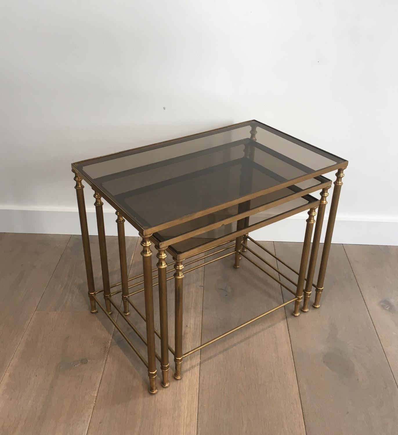 In the style of Maison Jansen. Set of 3 neoclassical brass nesting tables with fluted legs and smoked glass shelves, French, circa 1940.
