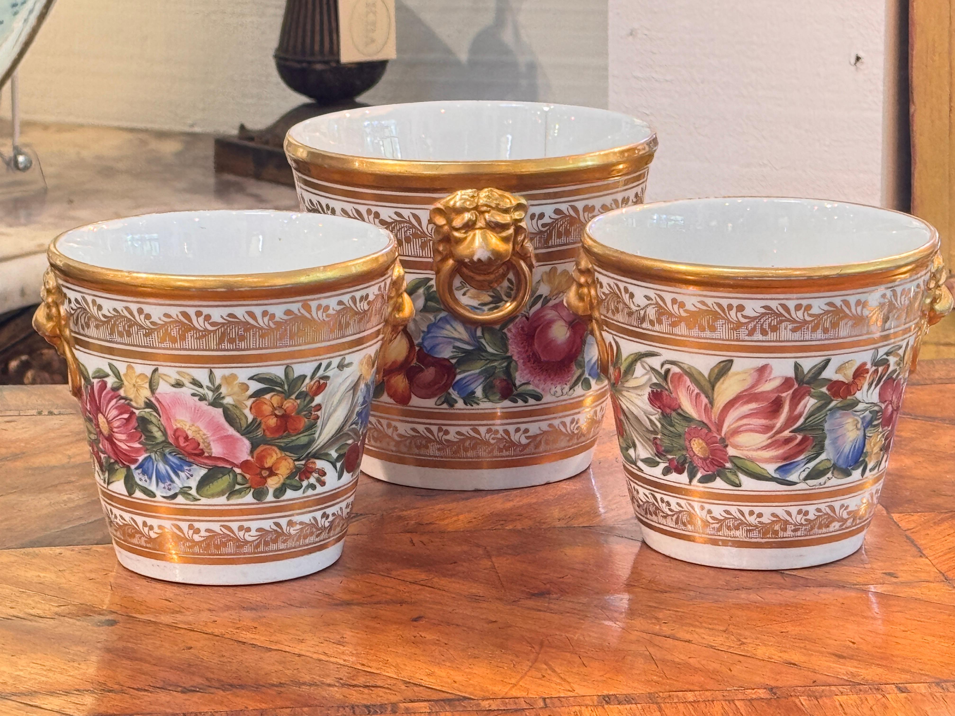 Ensemble de trois cache-pots en porcelaine, chacun richement peint à la main avec des guirlandes florales colorées et des détails dorés. Les bords et les bases sont ornés d'une fine bande dorée, tandis que les poignées en forme de masque de lion