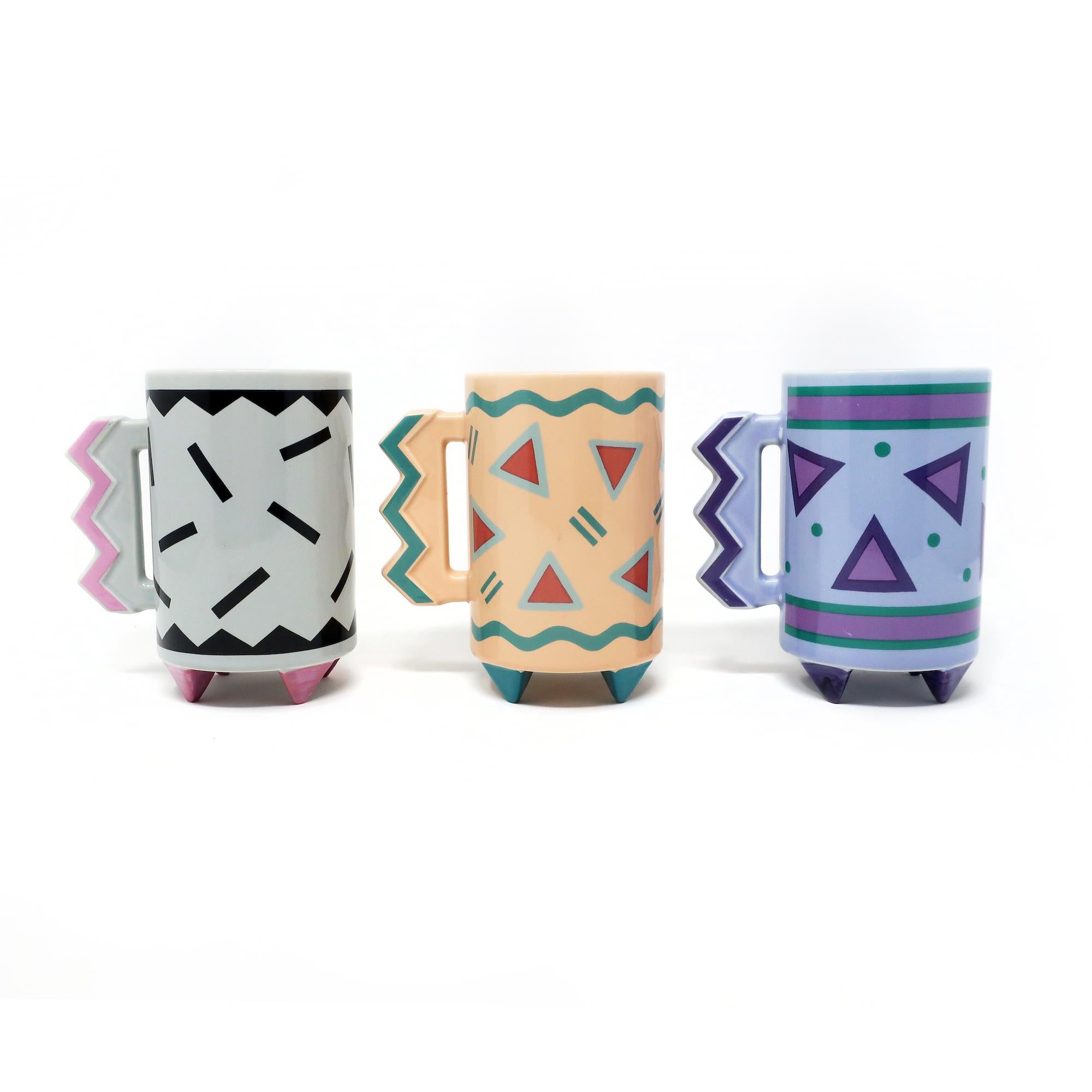 A super fun set of three postmodern mugs from 1989 made by Hallmark Cards and unquestionably influenced by the Memphis design movement. Each slim mug sits on four triangular feet and has a handle notched with triangles. One mug has a gray body with