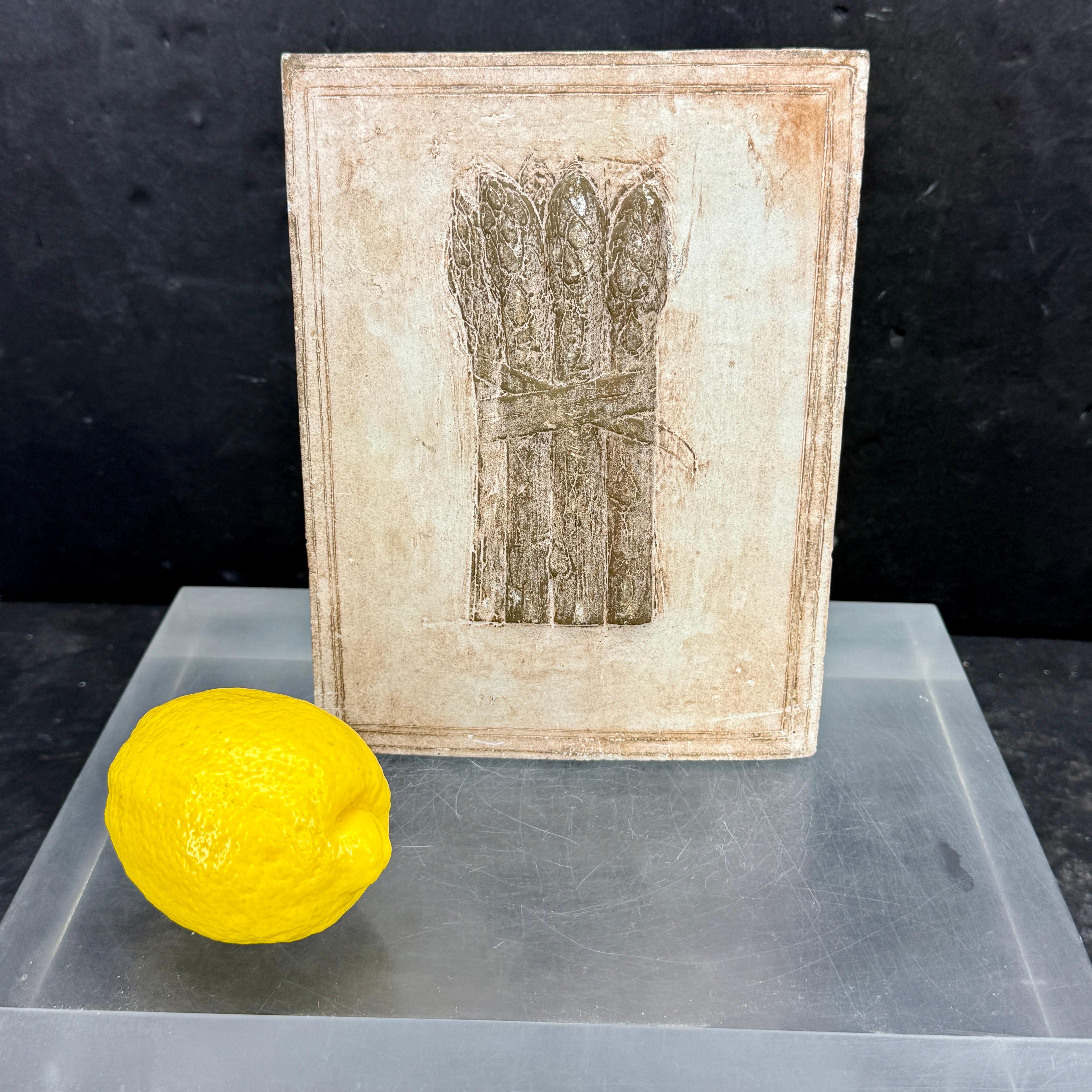 Encantador juego de tres azulejos de cerámica con una remolacha, un espárrago y una alcachofa. Los vemos en una estantería de una cocina de estilo europeo, rodeados de libros de cocina. Se pueden colgar en la pared. Firmado pero ilegible.