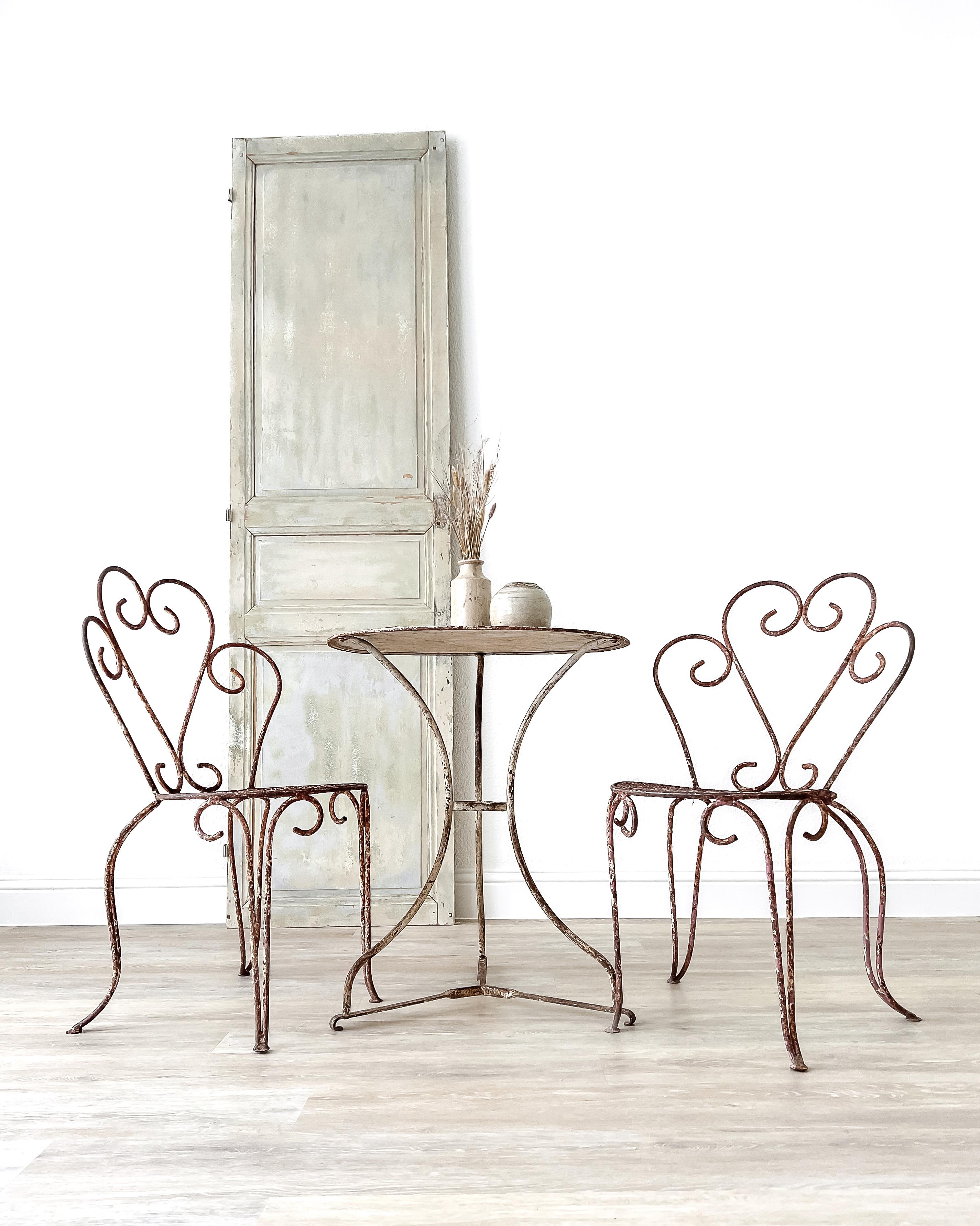 A chic and charming metal garden table with two matching chairs. Worn by the elements and showing myriad chippy paint layers, the set has acquired a wonderful patina full of unrivaled character. Having a simple design, the table is juxtaposed with a