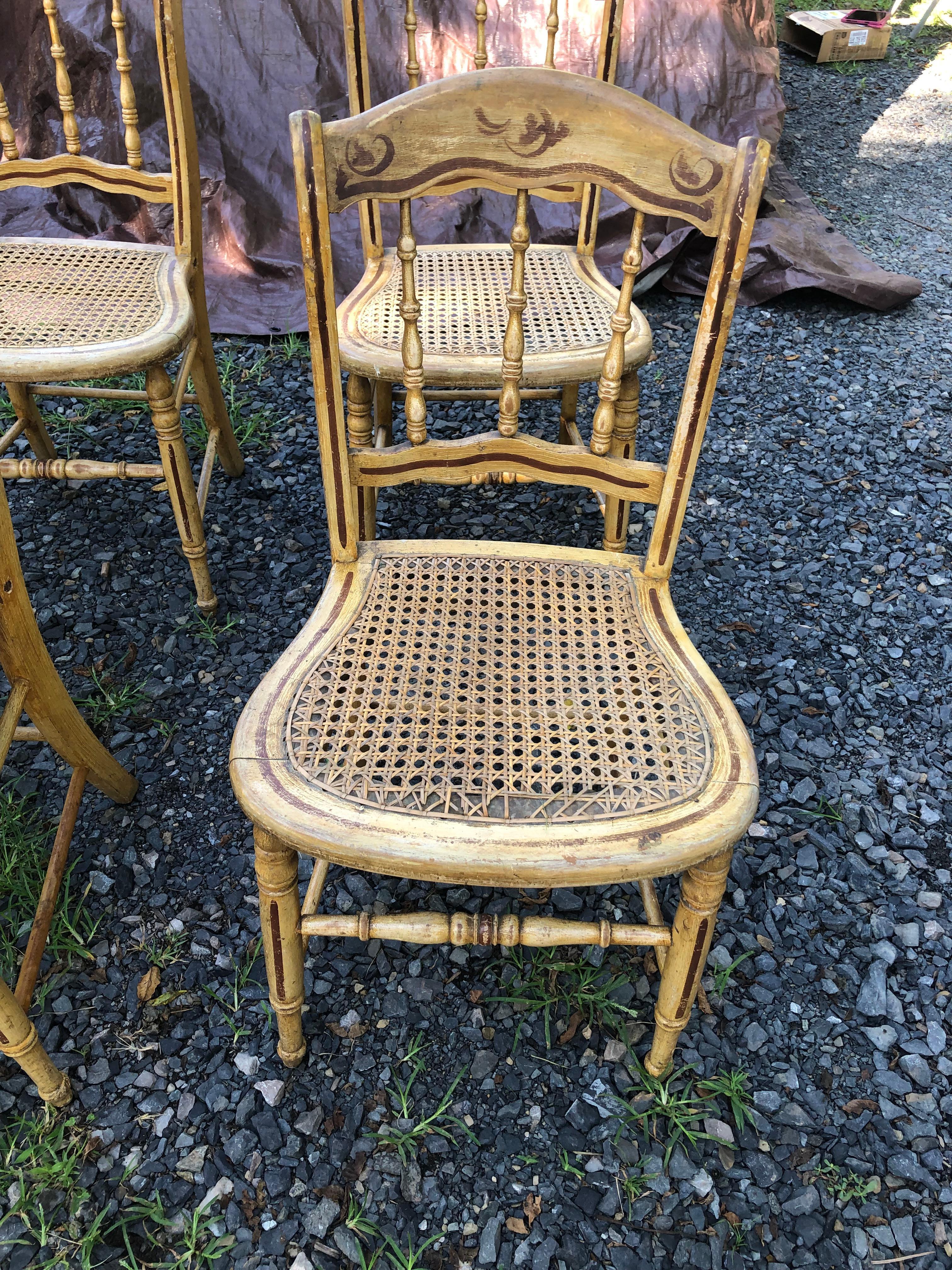 Ensemble de 4 charmantes chaises de salle à manger de fabrication Amish avec dossier en fuseau et assise cannée avec décoration peinte à la main.  Les chaises sont dans un état rustique, mais solides et robustes.  Un siège présente une usure visible