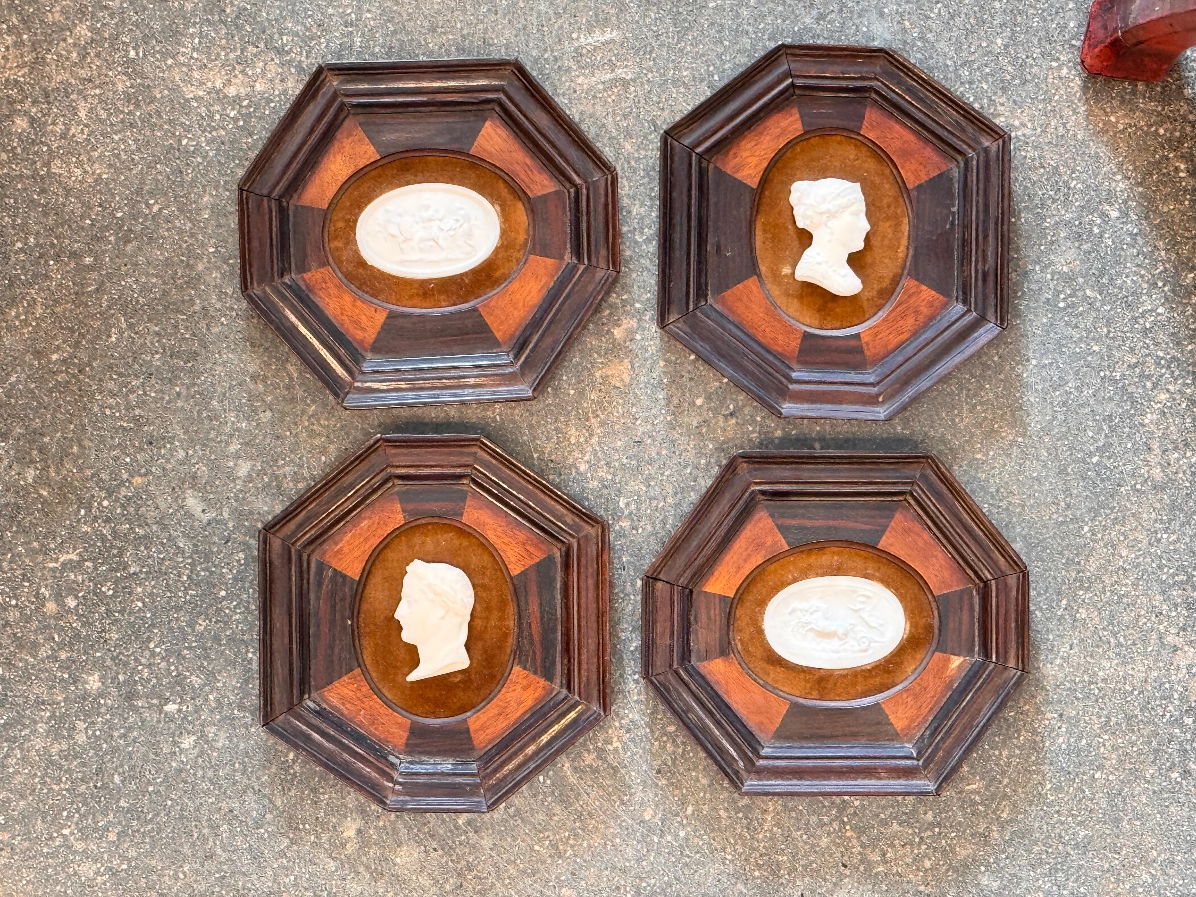 Satz von vier weißen Intaglien im klassischen Stil, die jeweils in einem kontrastierenden achteckigen Holzrahmen mit abwechselnden Platten montiert sind. Die Reliefs zeigen Profilporträts und mythologische Szenen und bieten einen zeitlosen