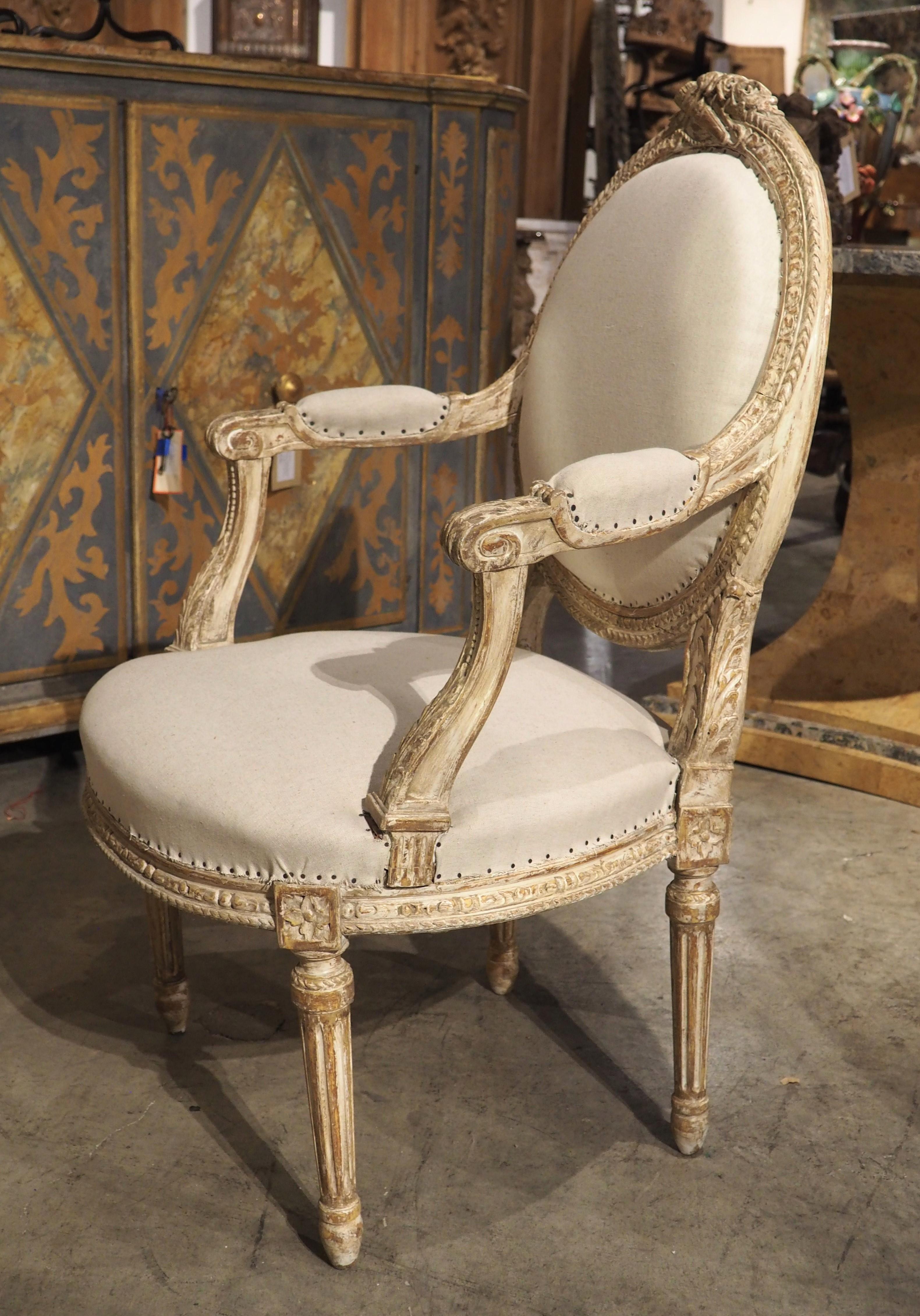 Dating to circa 1885, this set of four hand-carved parcel paint cabriolet armchairs are in the style of Louis XVI. The thick oval medallion backs (which have retained their original checkered canvas on the verso side) have been adorned with a trophy
