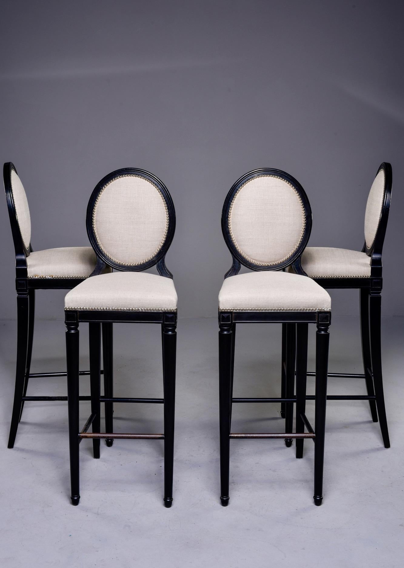 Found in England, this set of four bar height stools feature beech frames in a black finish with classic oval backs. Upholstery is natural colored linen with silver tone nail head trim. There are water spots and some rust marks from nail head tacks