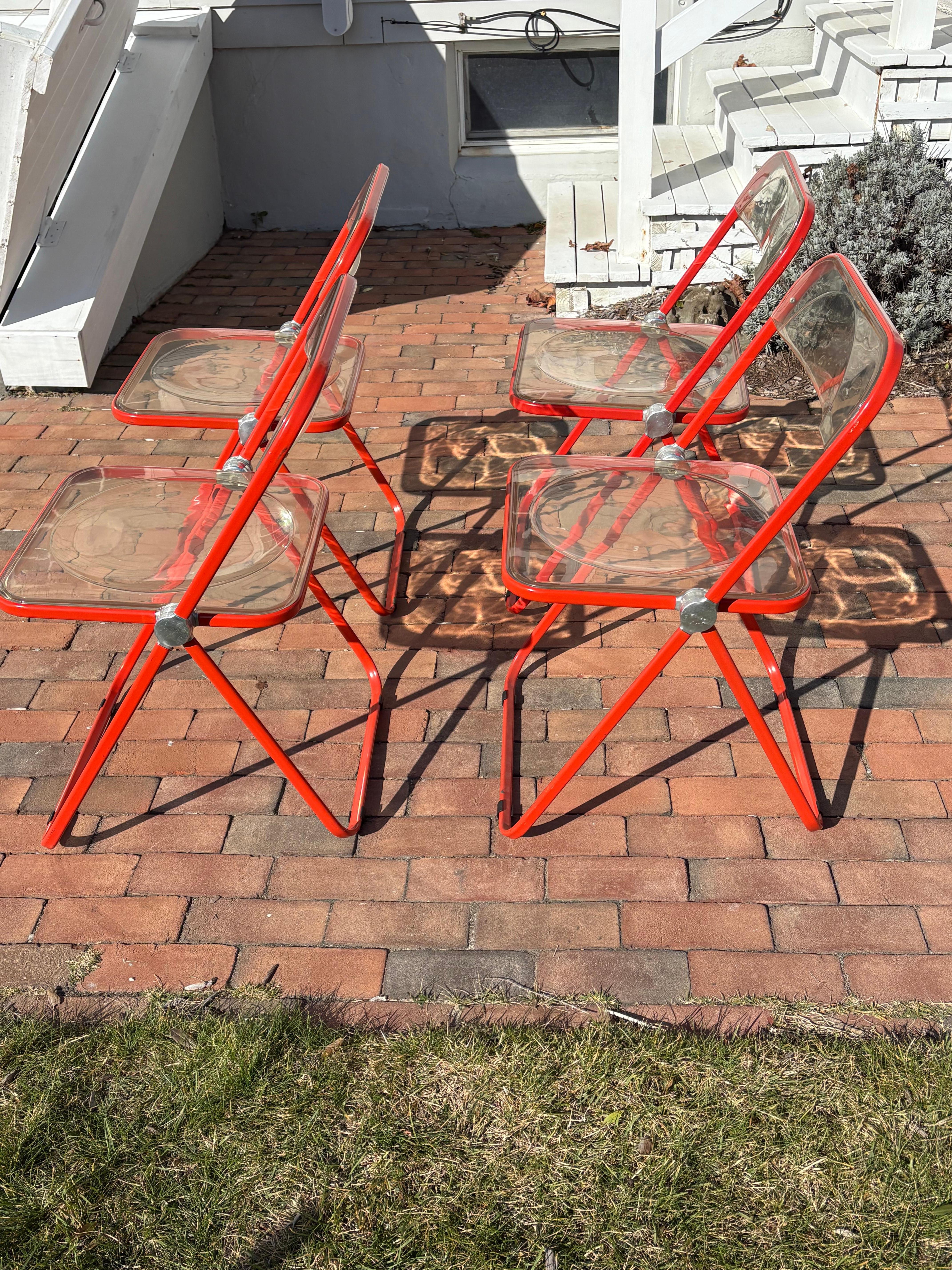 Ensemble de quatre sièges et dossiers en lucite claire des années 1970 avec des cadres en aluminium orange. Ces chaises pliantes Plia ont été conçues par Giancarlo Piretti pour Castelli. Fait en Italie. La chaise Plia est un classique du design au