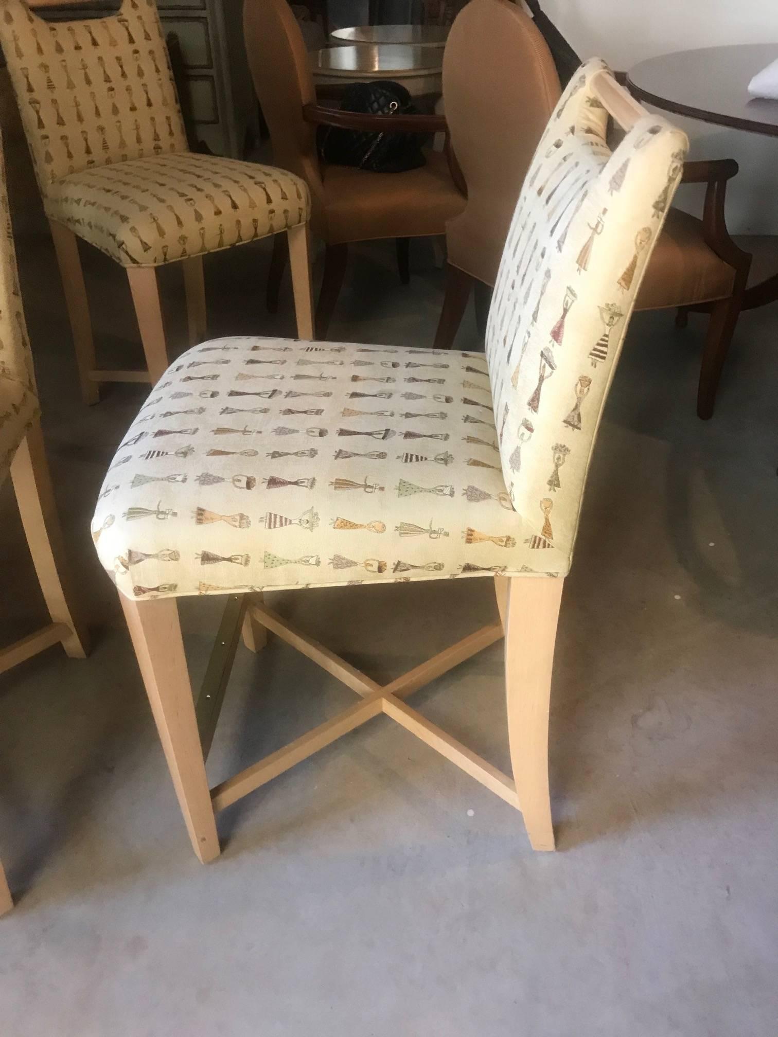 A set of four counter height very stylish chairs having a chic Italian look with light wood angular legs and dowel at the top, upholstered in a fun beige fabric patterned with fashionable ladies in different attire. Rich looking brass plates at the