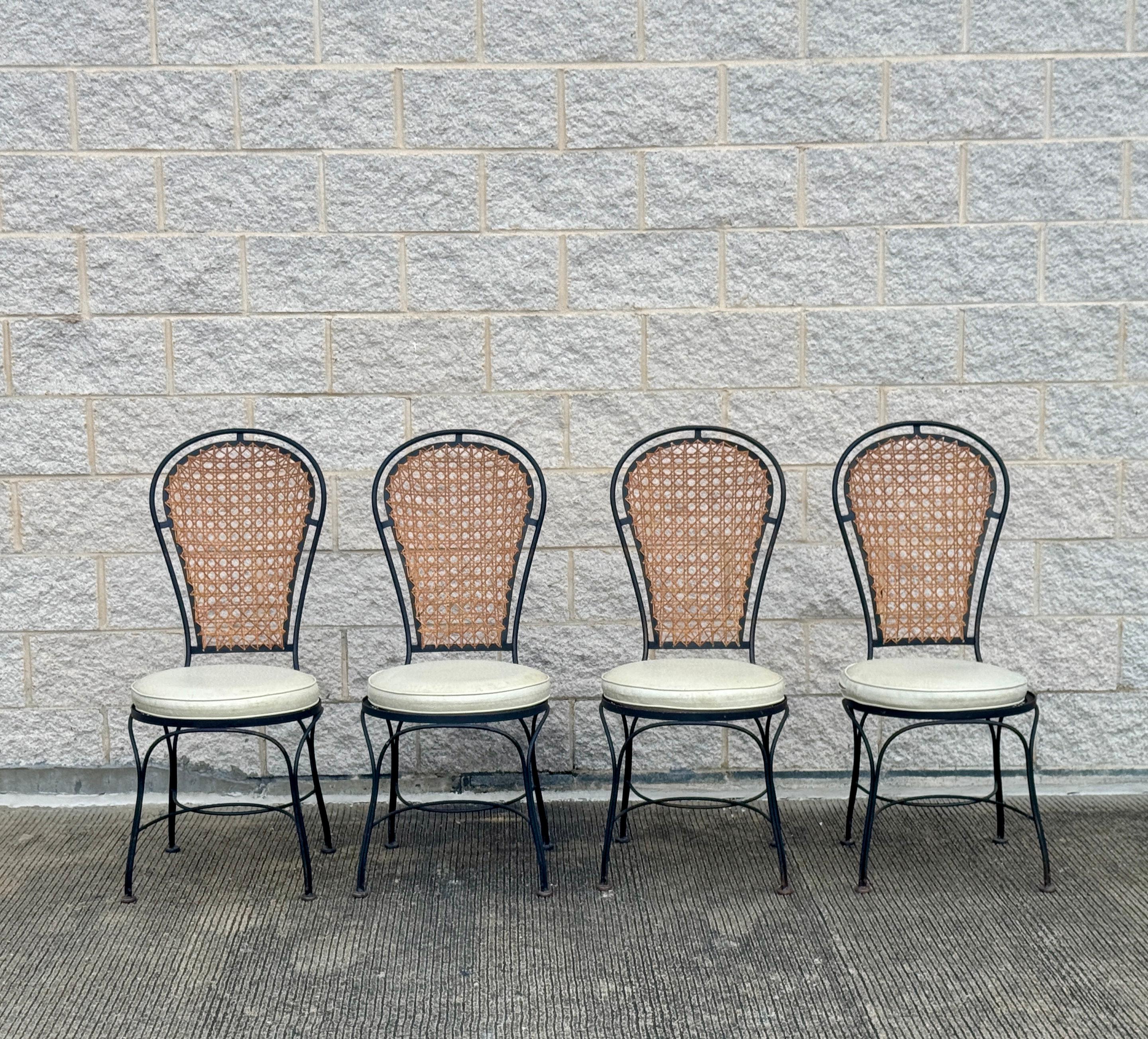 Set di quattro sedie da pranzo vintage con struttura in ferro battuto nero e schienale curvo, inserti in canna naturale e cuscini di seduta rotondi imbottiti in ecopelle bianco sporco. I cuscini sono rimovibili e sembrano essere originali. Non è