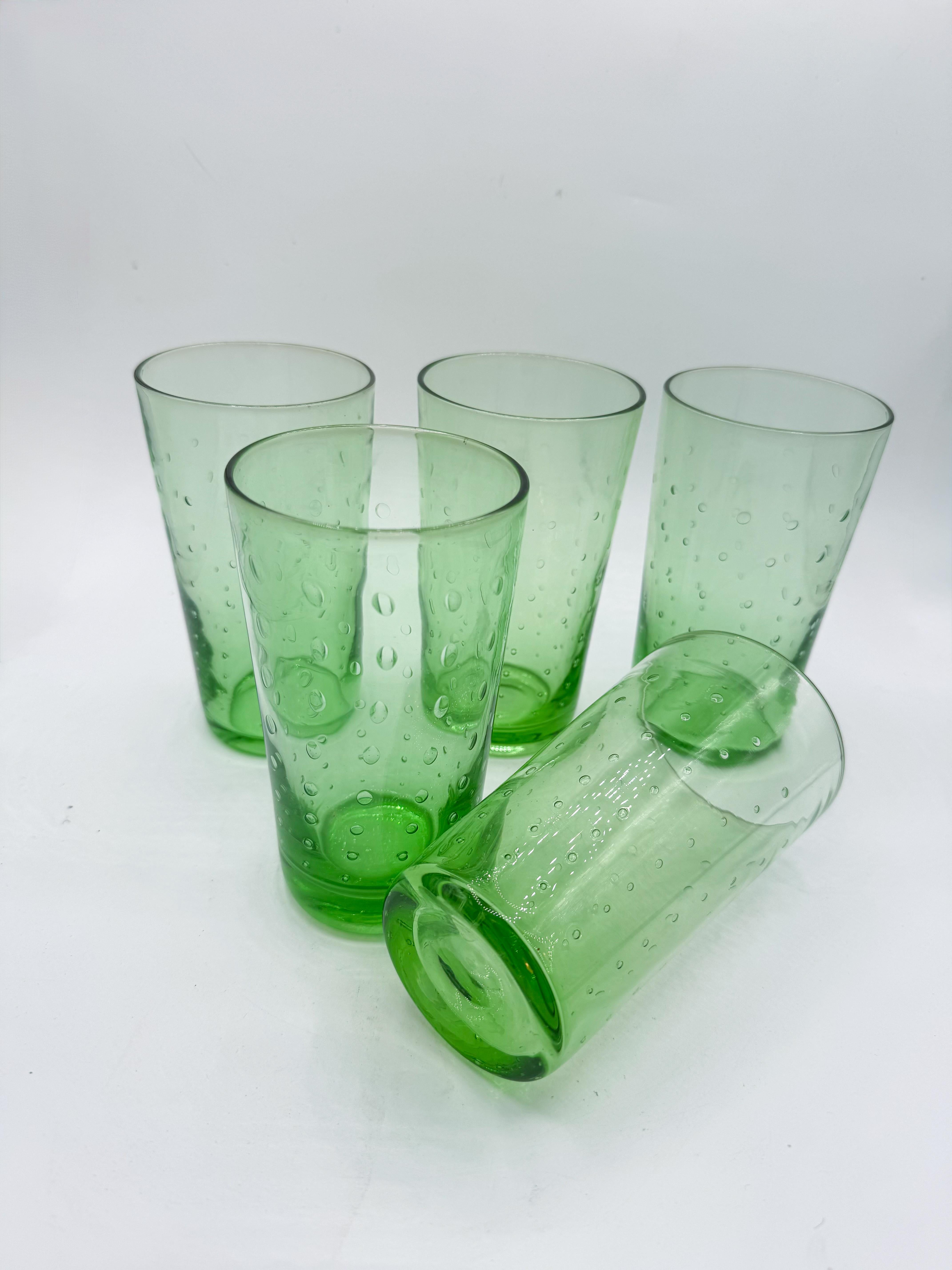A charming set of five hand-blown green glass tumblers featuring a lively seeded bubble design. Each glass showcases unique variations in bubble placement, a hallmark of artisanal craftsmanship that adds character and individuality to the set. The