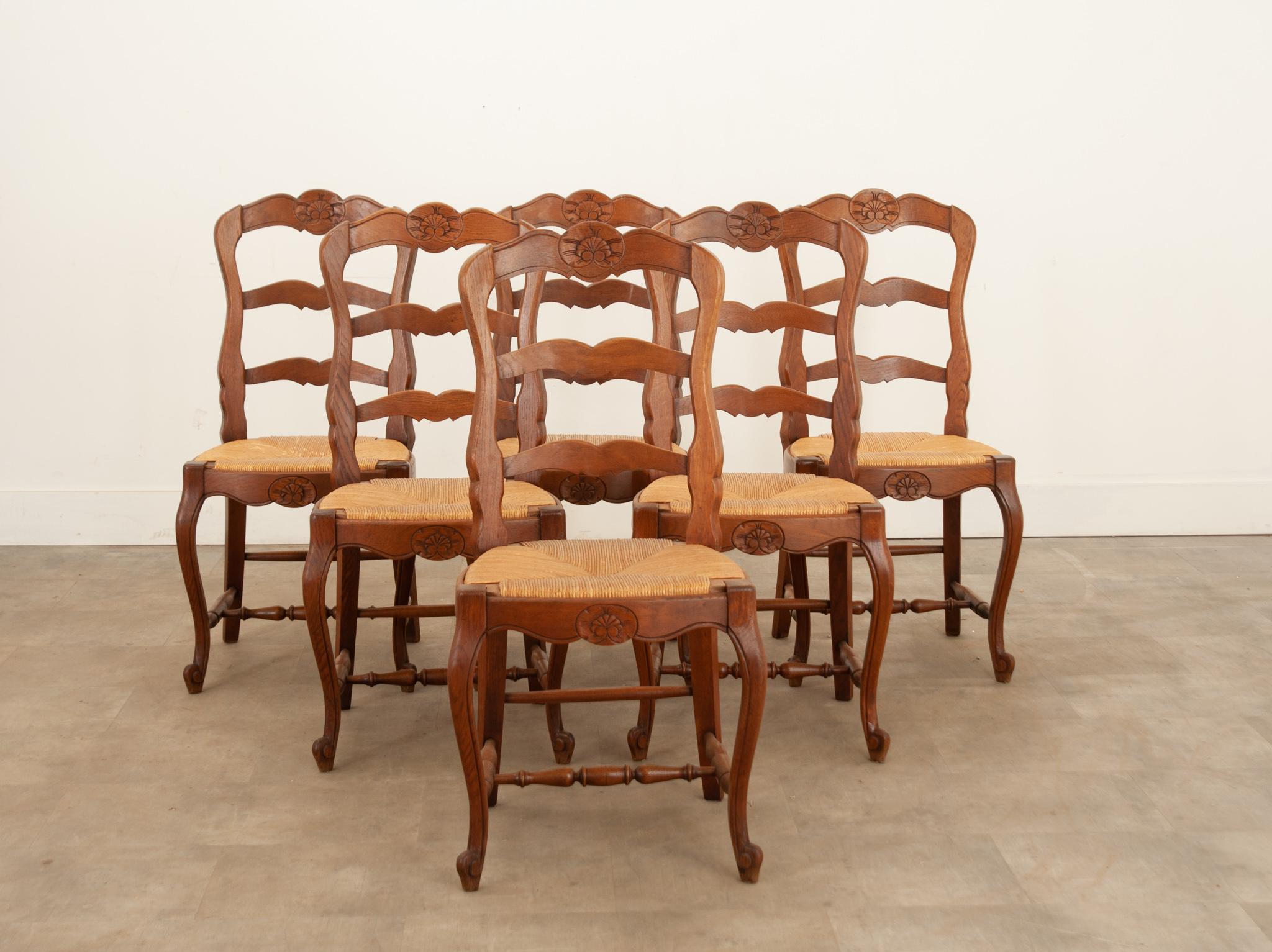 A charming set of six French oak ladder back chairs made in the 1940s. A beautiful carved shell cartouche graces the chairs’ top and front rails. The rush seats are in excellent antique condition and have a height of 18 1/2?. The seats are