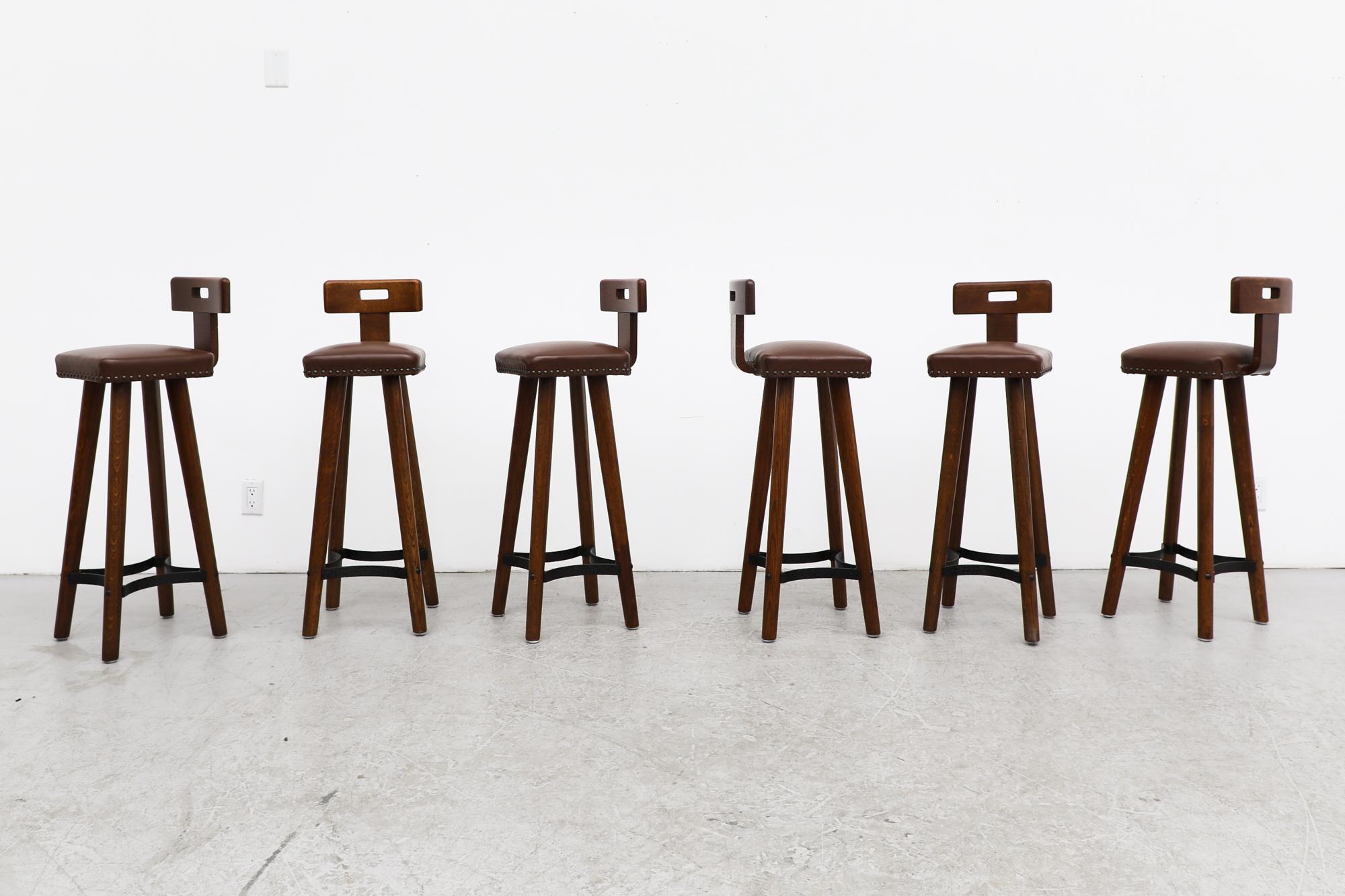This mid century set of handsome, heavy oak T-Back bar stools inspired by the designs of French master Pierre Chapo have a small handle cut-out on the backrest and feature a Brutalist iron foot rest. The seats are made leather upholstered with
