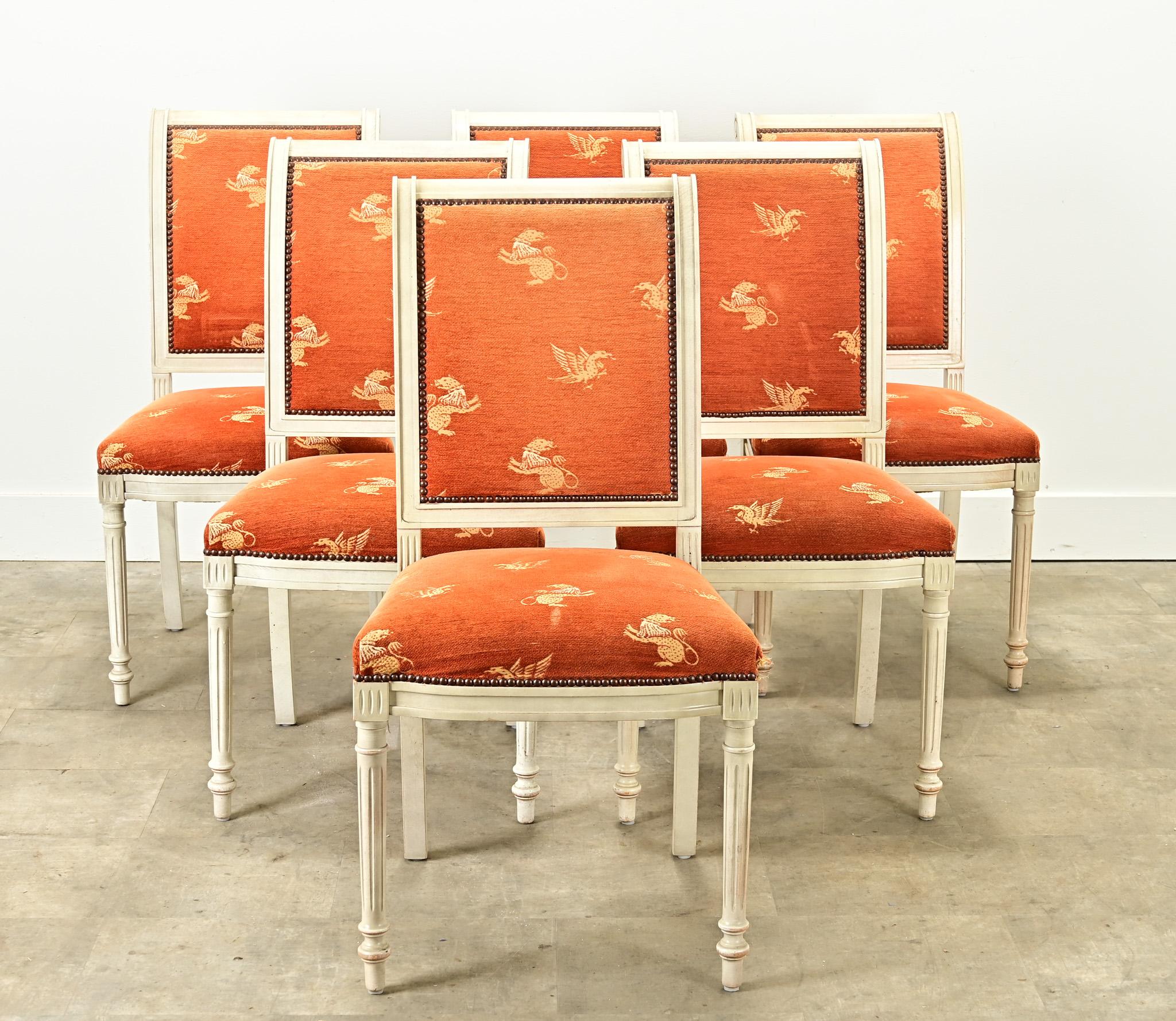 A vintage set of six Louis XVI style upholstered dining chairs with painted frames. These comfortable dining chairs have a worn bright orange fabric with griffin motifs secured by brass nailheads. The carved and painted frames are sturdy and painted