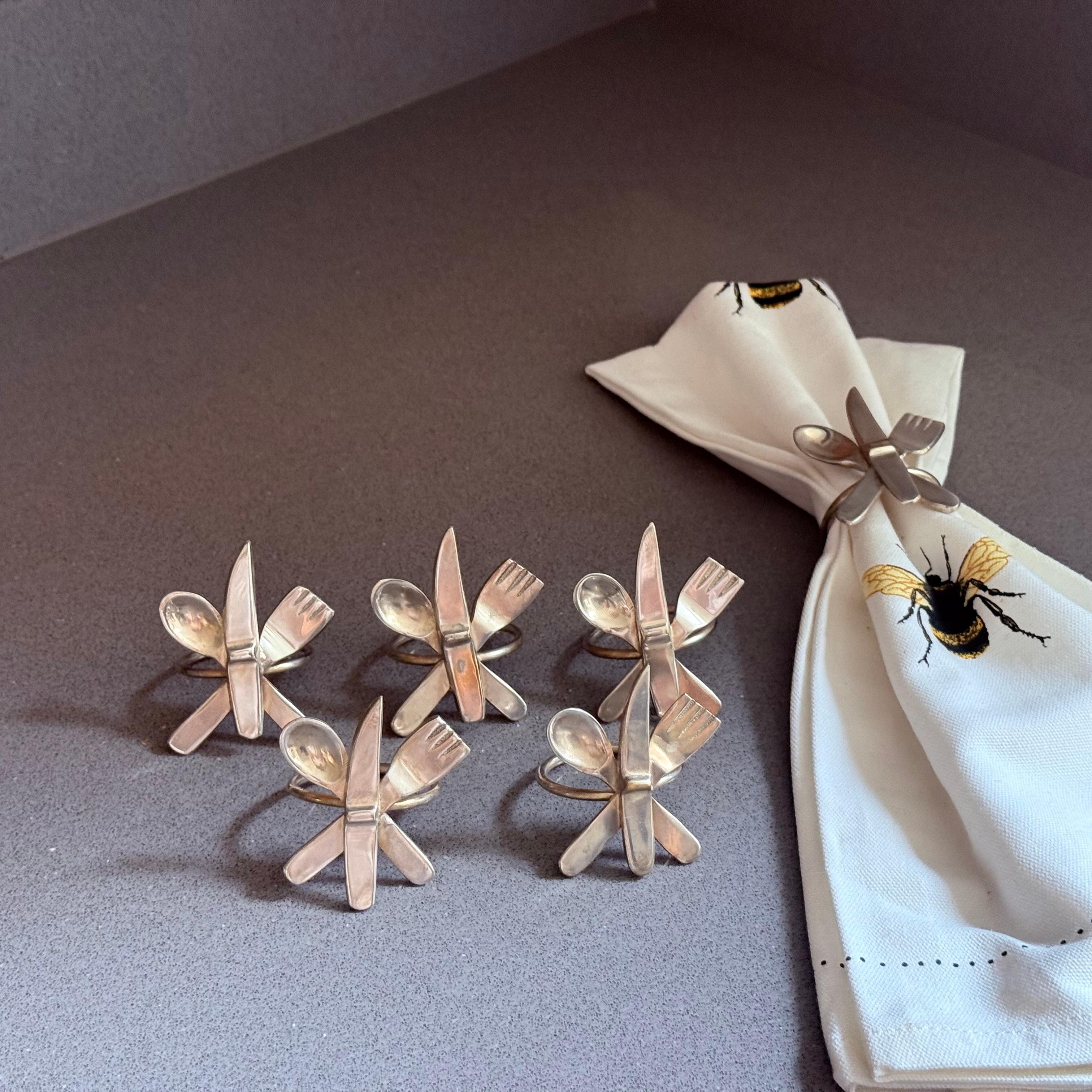 Ein verspieltes und zugleich elegantes Set aus sechs versilberten Serviettenringen im Vintage-Stil, die jeweils mit einem charmanten gekreuzten Besteckmotiv aus Gabel, Messer und Löffel versehen sind. Diese einzigartigen Tischaccessoires bringen