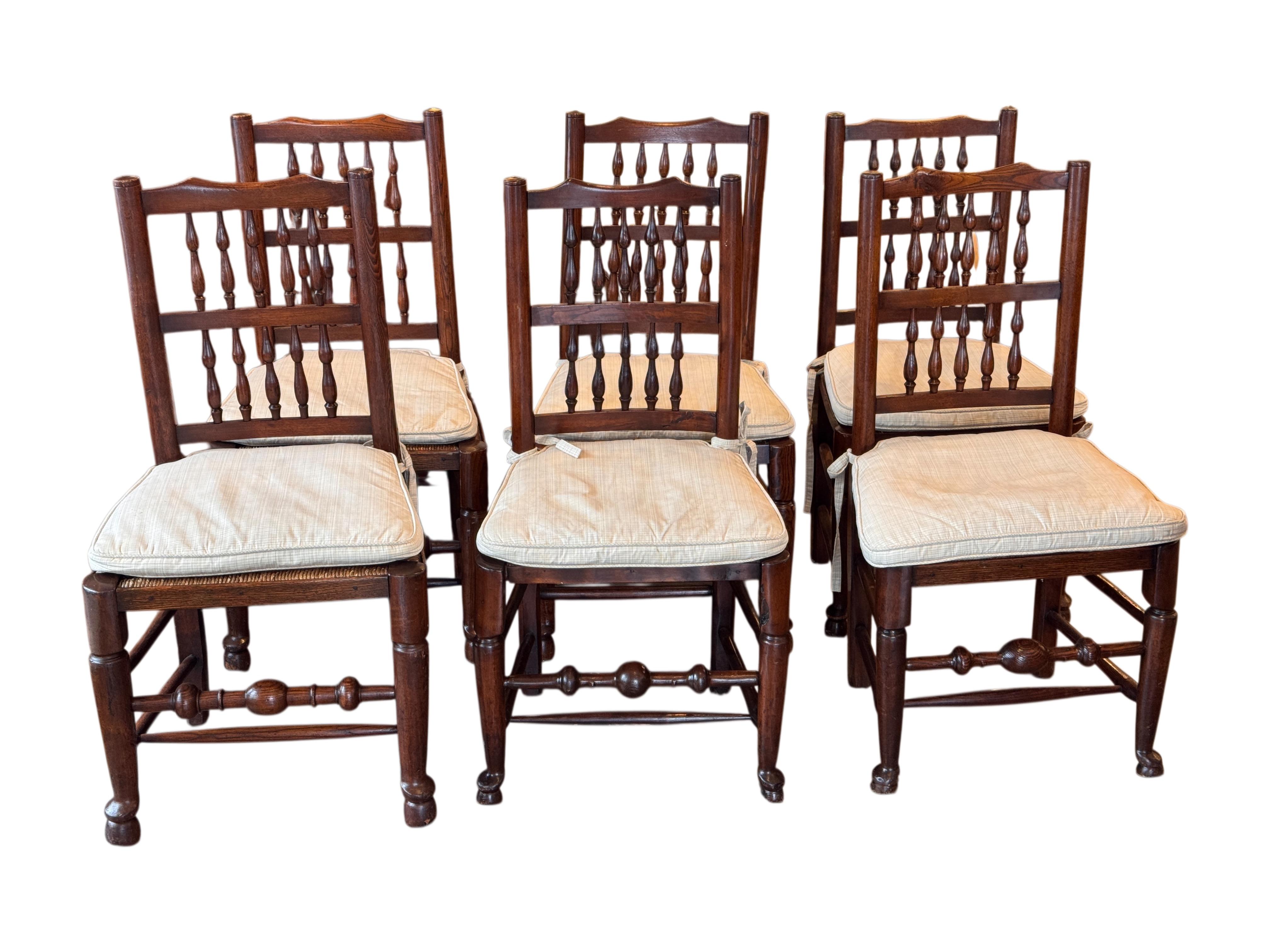 Set of six spindle-back chairs with turned back posts, shaped crest rails, and multiple tapered spindles. The seats are woven rush, and the frames feature turned legs and stretchers, including a baluster-form front stretcher. Cushions are shown