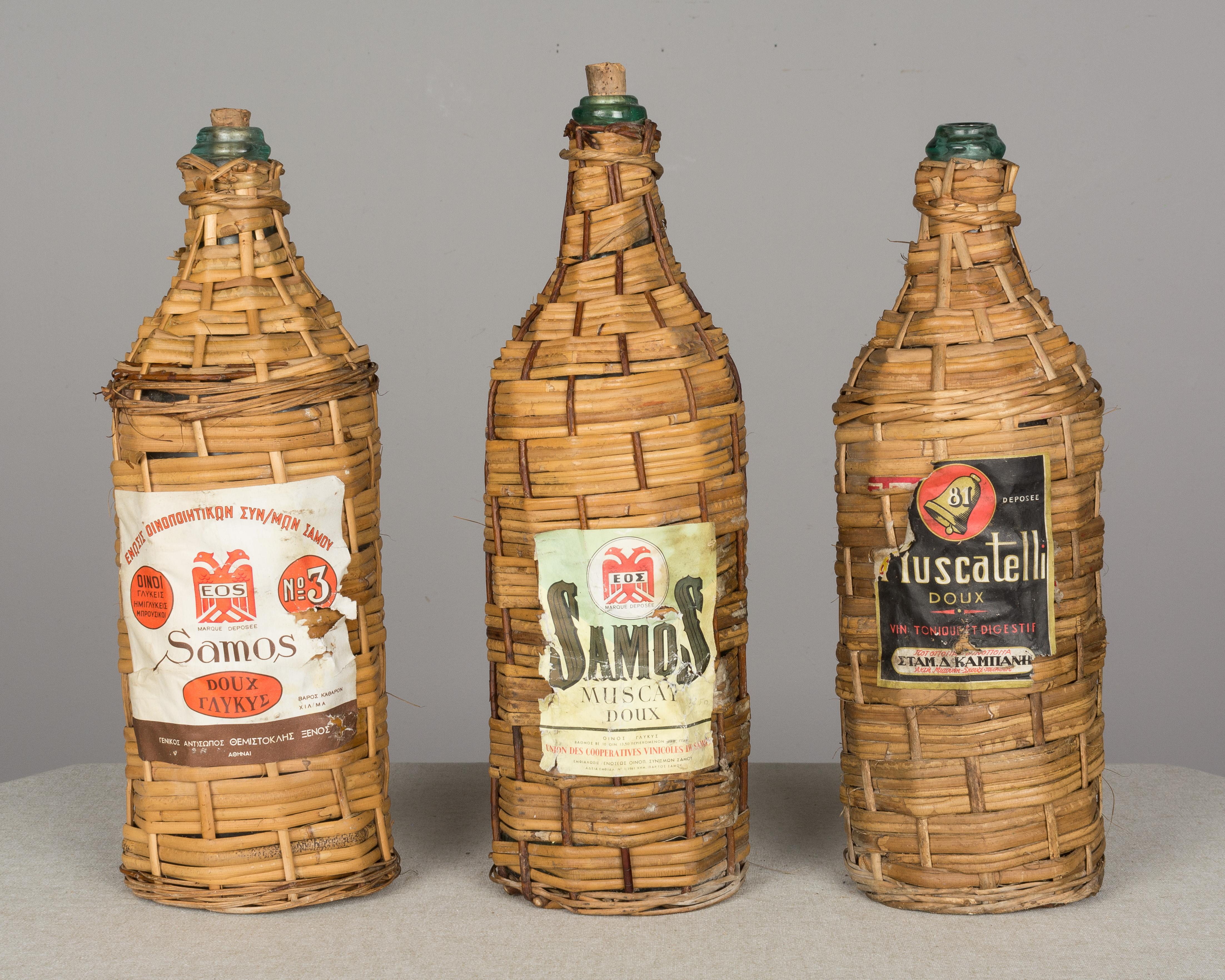 A set of six vintage wicker-wrapped glass bottles with handles. Original paper wine labels on three, two with corks. Smallest bottle with double handles is more tightly woven. Purchased along with a midcentury rattan bar from the French