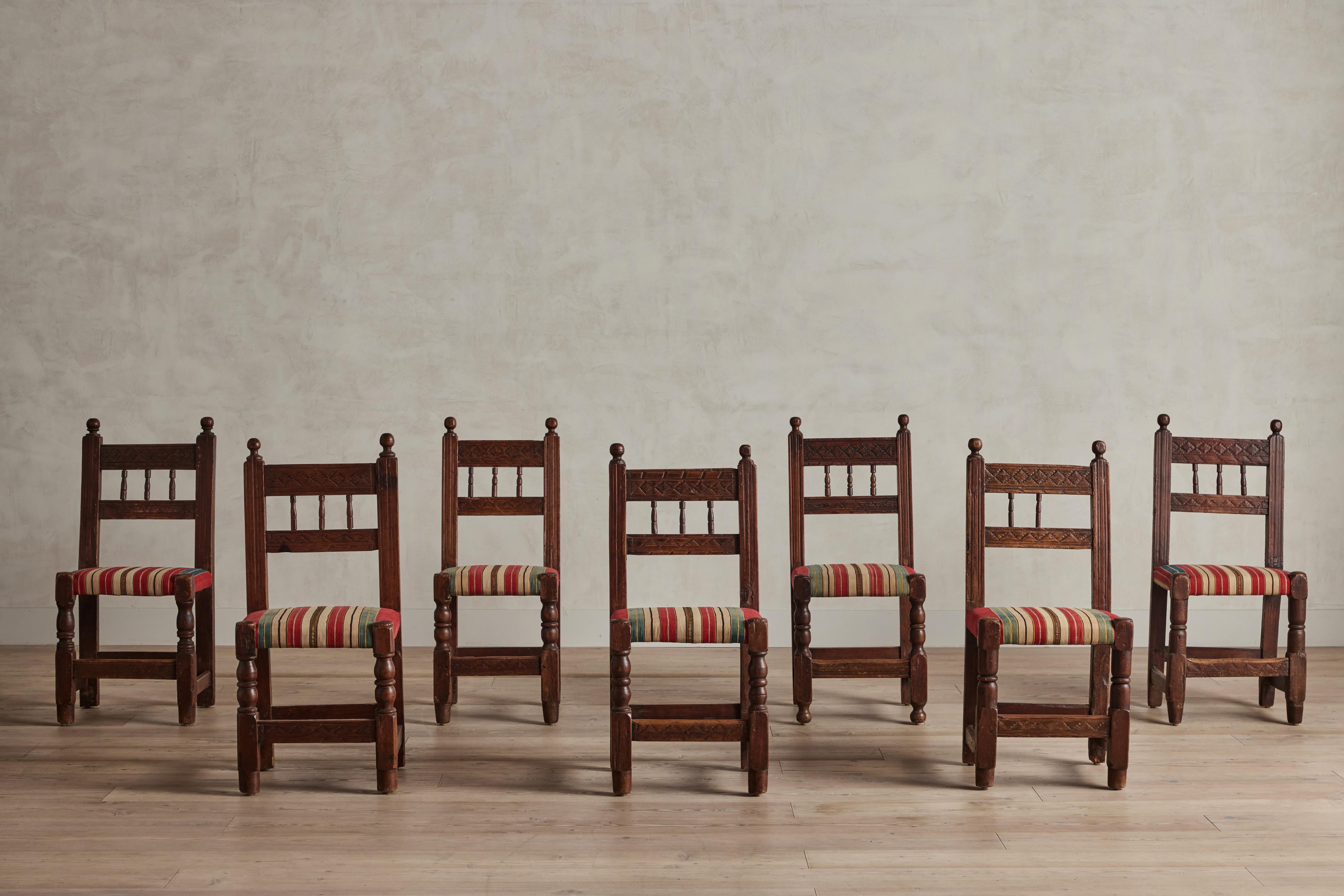  Set of seven carved wood dining chairs from Spain circa 1920. Newly upholstered in a striped French cotton and silk blend woven textile. There is wear throughout on the wood and fabric that is consistent with age and use. One chair is missing a