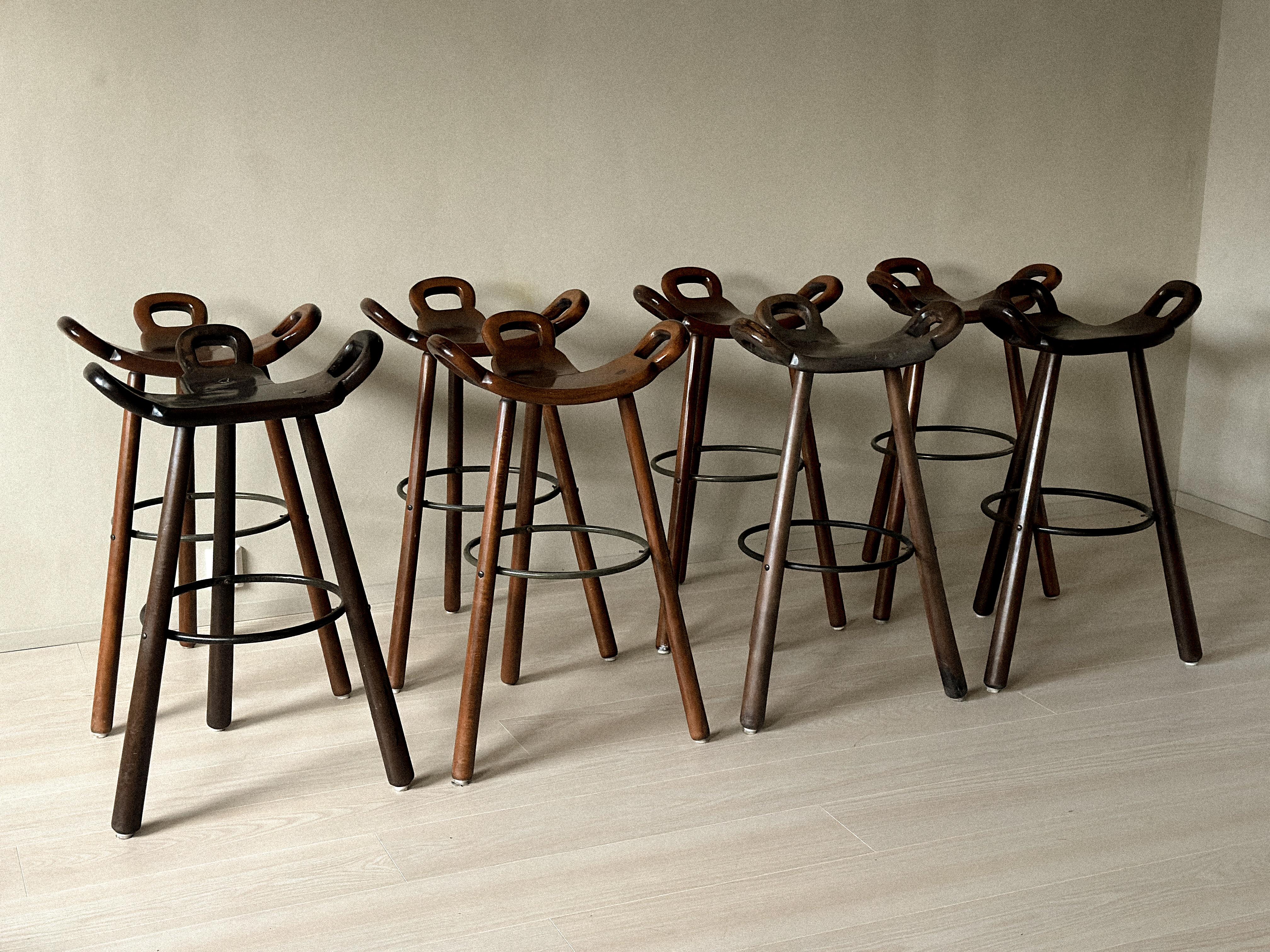 Set of 8 Brutalist-inspired bar stools in stained beech and steel, Europe, circa 1970s. The sculpted, saddle-shaped seats are carved with a gentle curvature that embraces the body, while cut-out handles on either side serve as practical handholds.