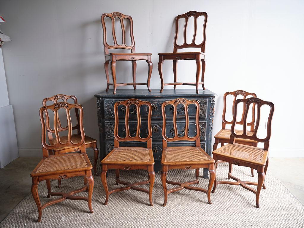 This is a lovely set of 8 English cane seat dining chairs. They have wood carved features, cabriole legs, and cane seats. The wood coloring is a patina light brown, and each chair has a beautiful wood carved detail across the top of the back of the