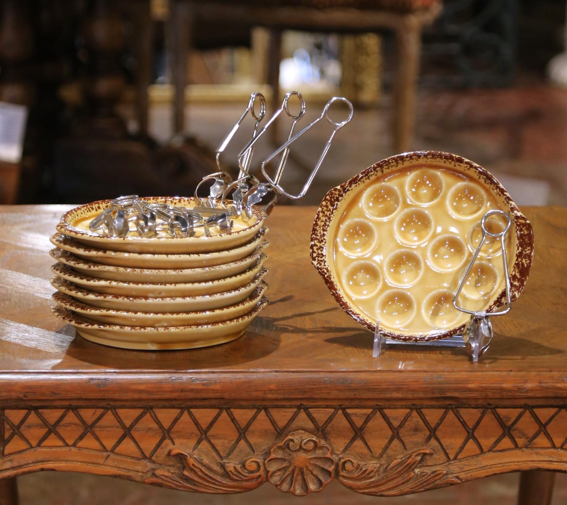 Crafted in France by Gien circa 1920, this elegant service set includes eight porcelain escargot plates and eight silver-plated spoons. Each plate, measuring 8.25 inches in diameter by 7.5 inches across the handles, features twelve molded wells for