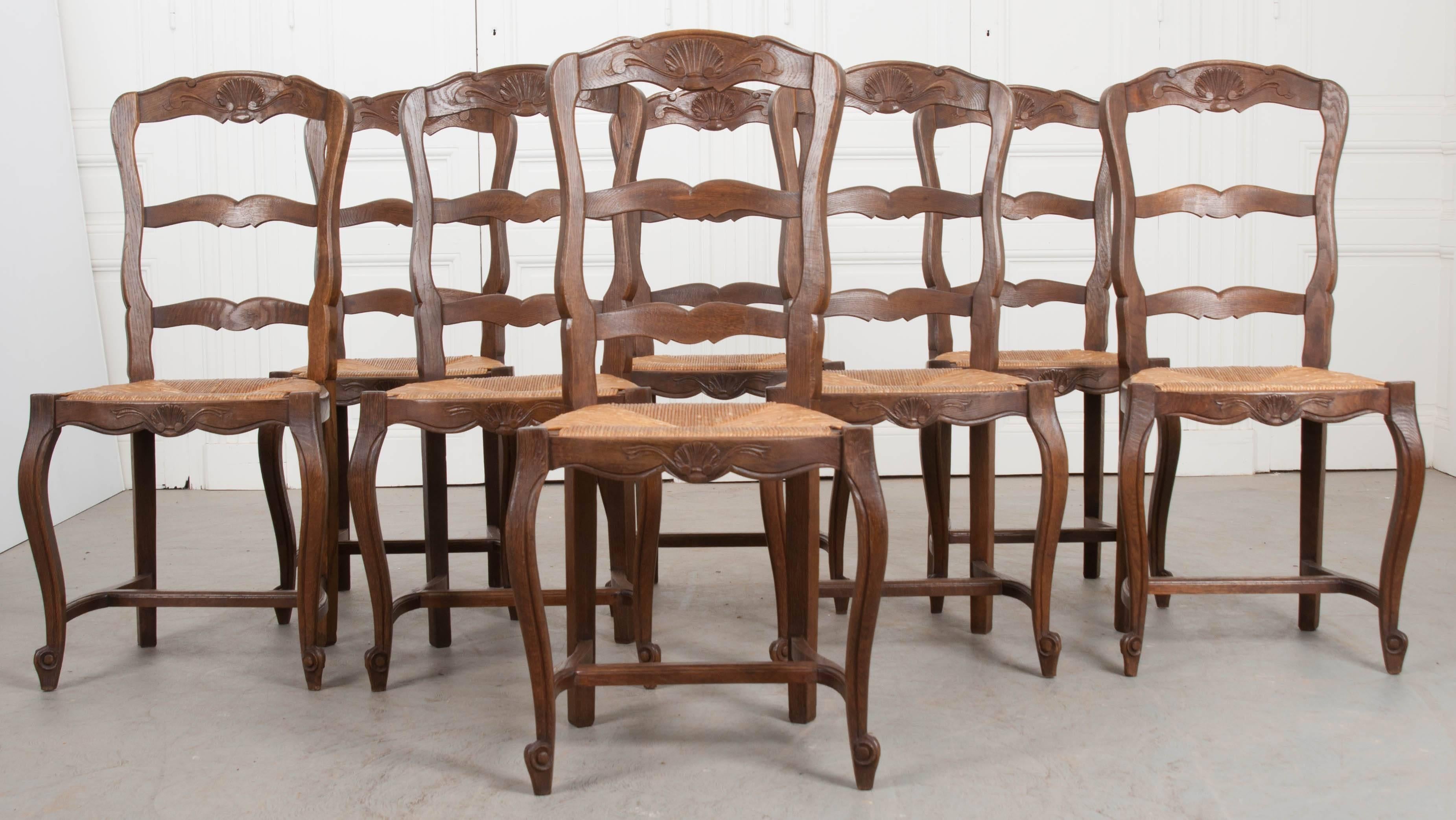 A set of eight Provincial style oak ladder back dining chairs, with rush seats, from France. This set was made in the 1930s and are in wonderful condition. The top and shaped front rails are each emblazoned with beautifully carved shell cartouches.