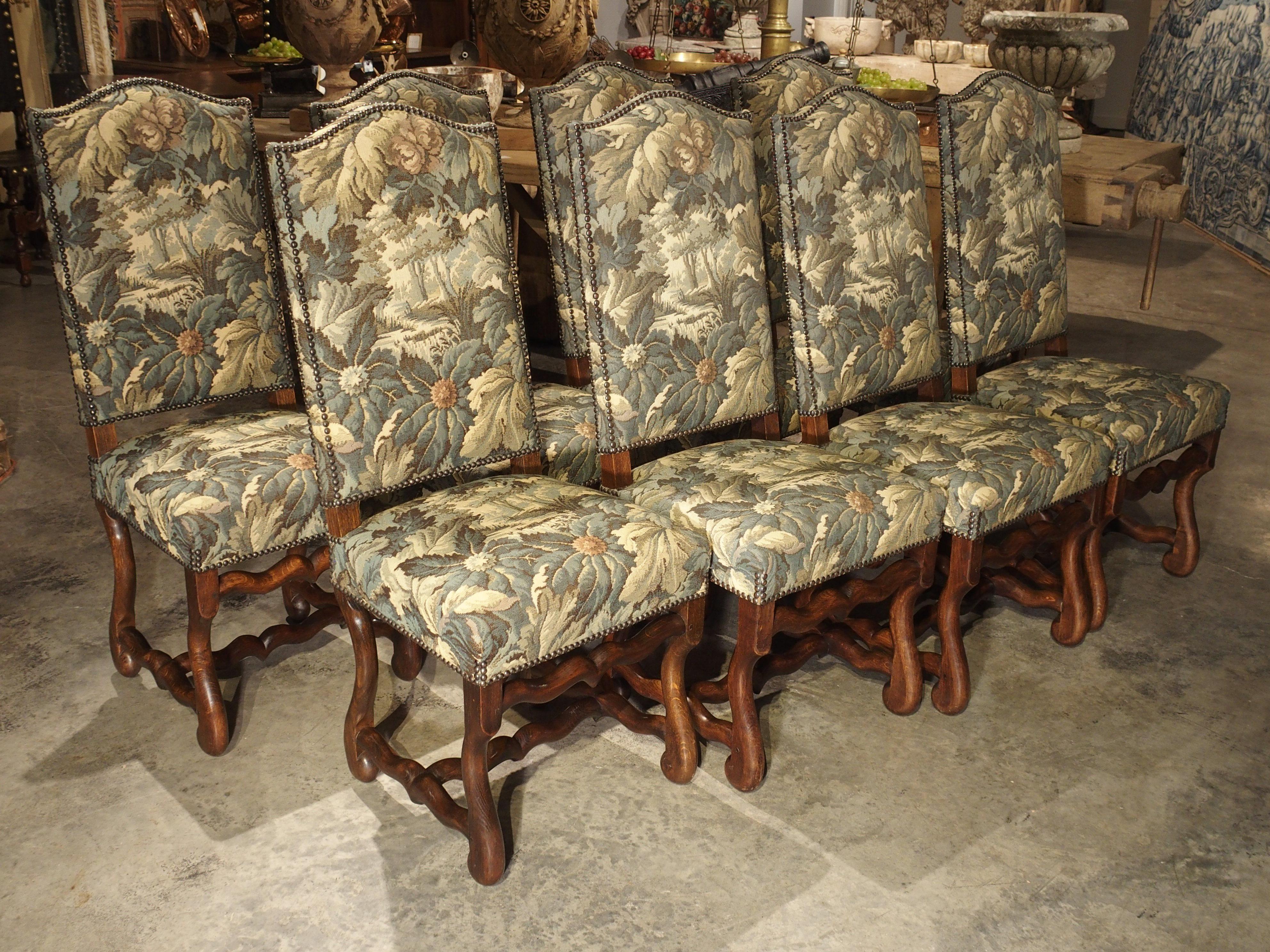 This classic group of 8 French Os de Mouton oak dining chairs have been upholstered with machine made verdure tapestry. The tapestries are attached to the frames with hundreds of brass nailheads, and the oak frames are carved in a style which