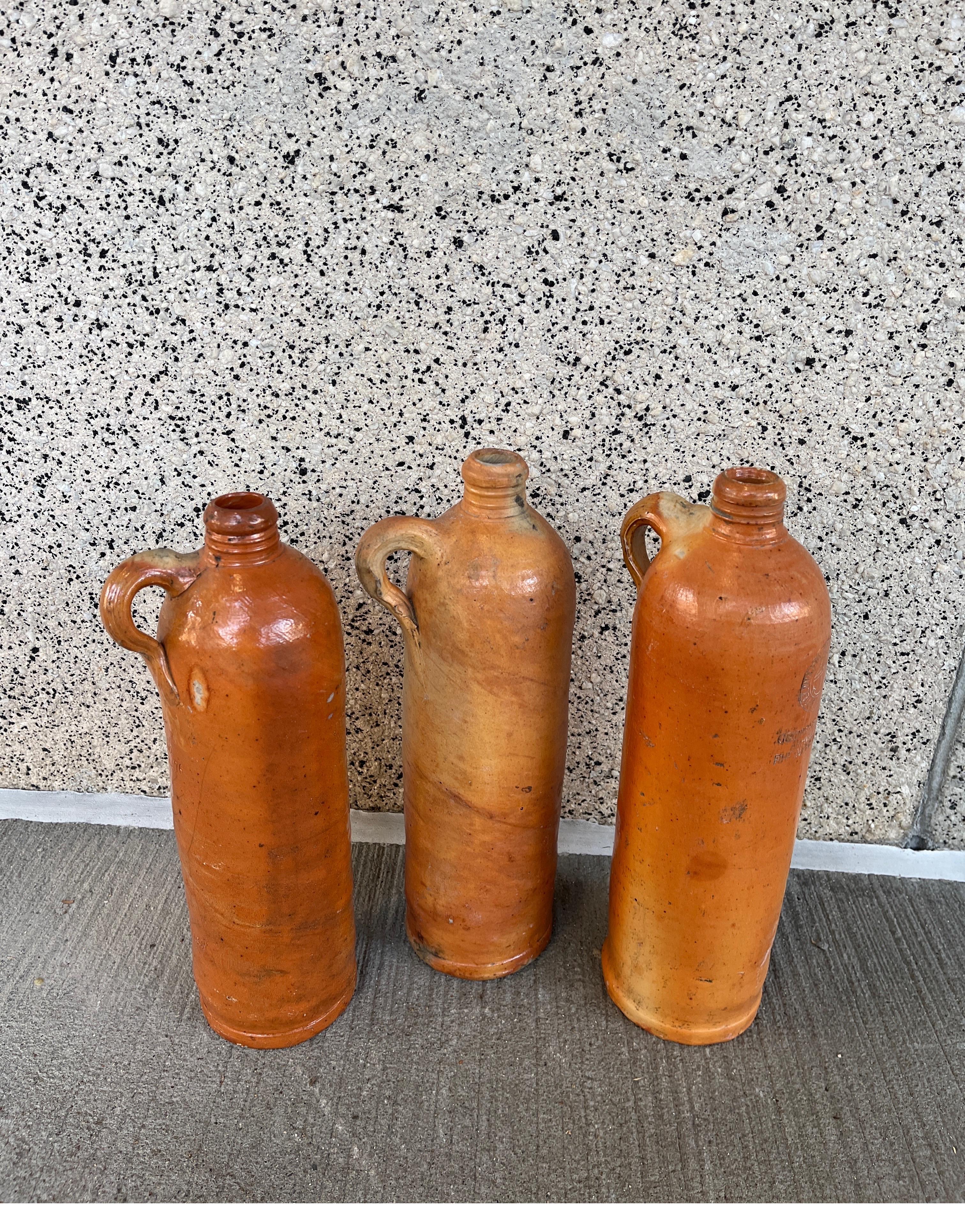 A set of three antique stoneware gin bottles from Germany. Each one has the impressed name of the maker. Gracefully shaped, with small thumb handles, these tall jars originally contained gin or other alcoholic beverages. Their soft colors,