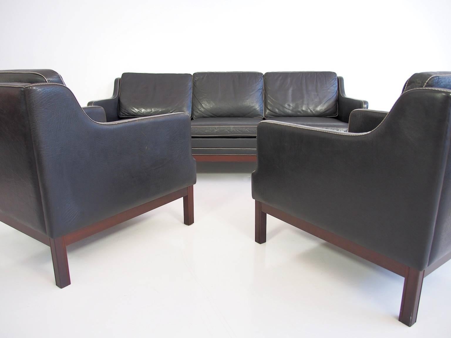 Black buffalo leather sofa and two accompanying easy chairs. Danish furniture producer. The three-seat sofa and armchairs are upholstered in black buffalo leather with white stitching. Mahogany-stained beech base. Beautiful natural patina. The width